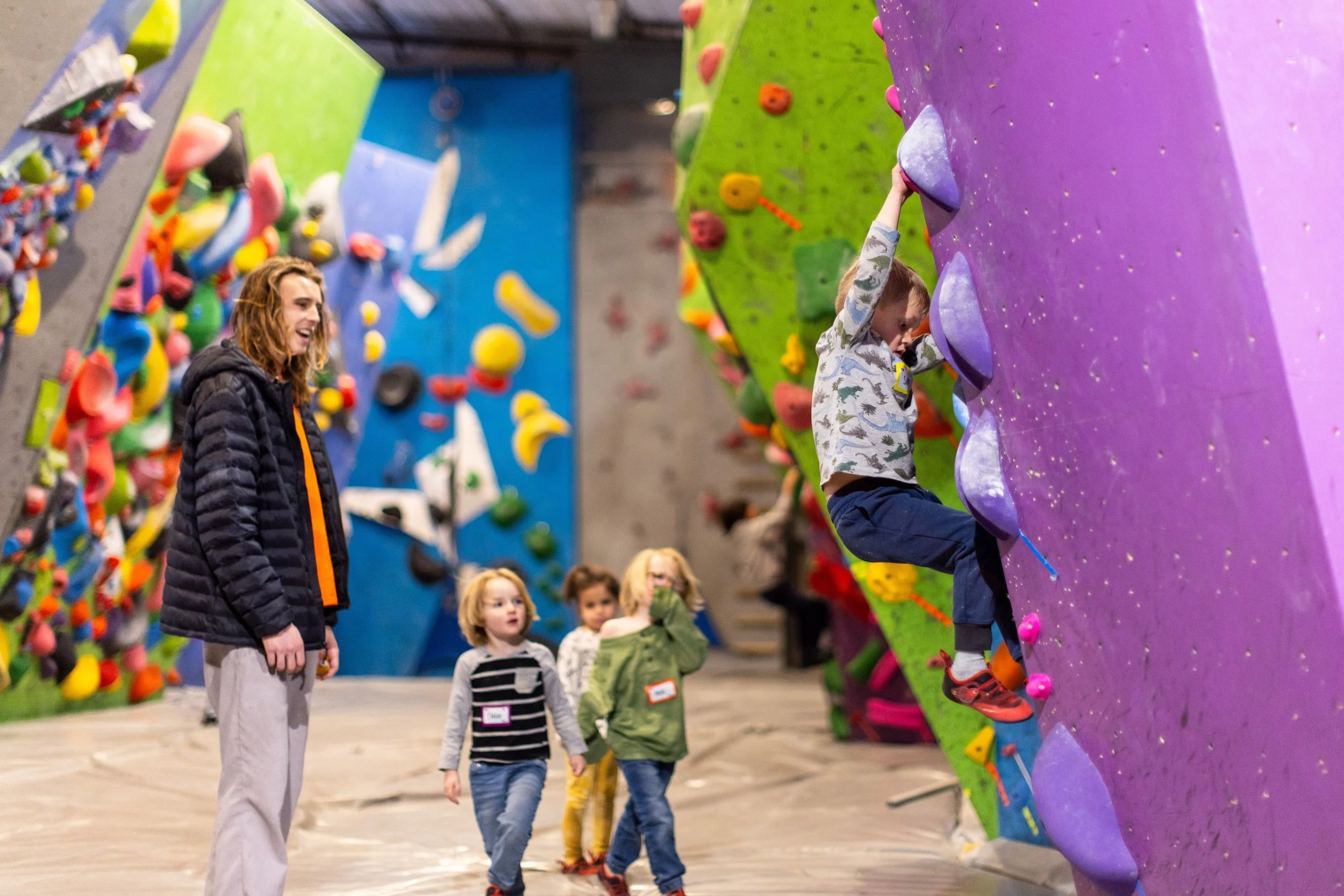 Young Hot Shots Prep climbers walking through the gym while a teammate climbs a colorful wall at ABC Kids Climbing