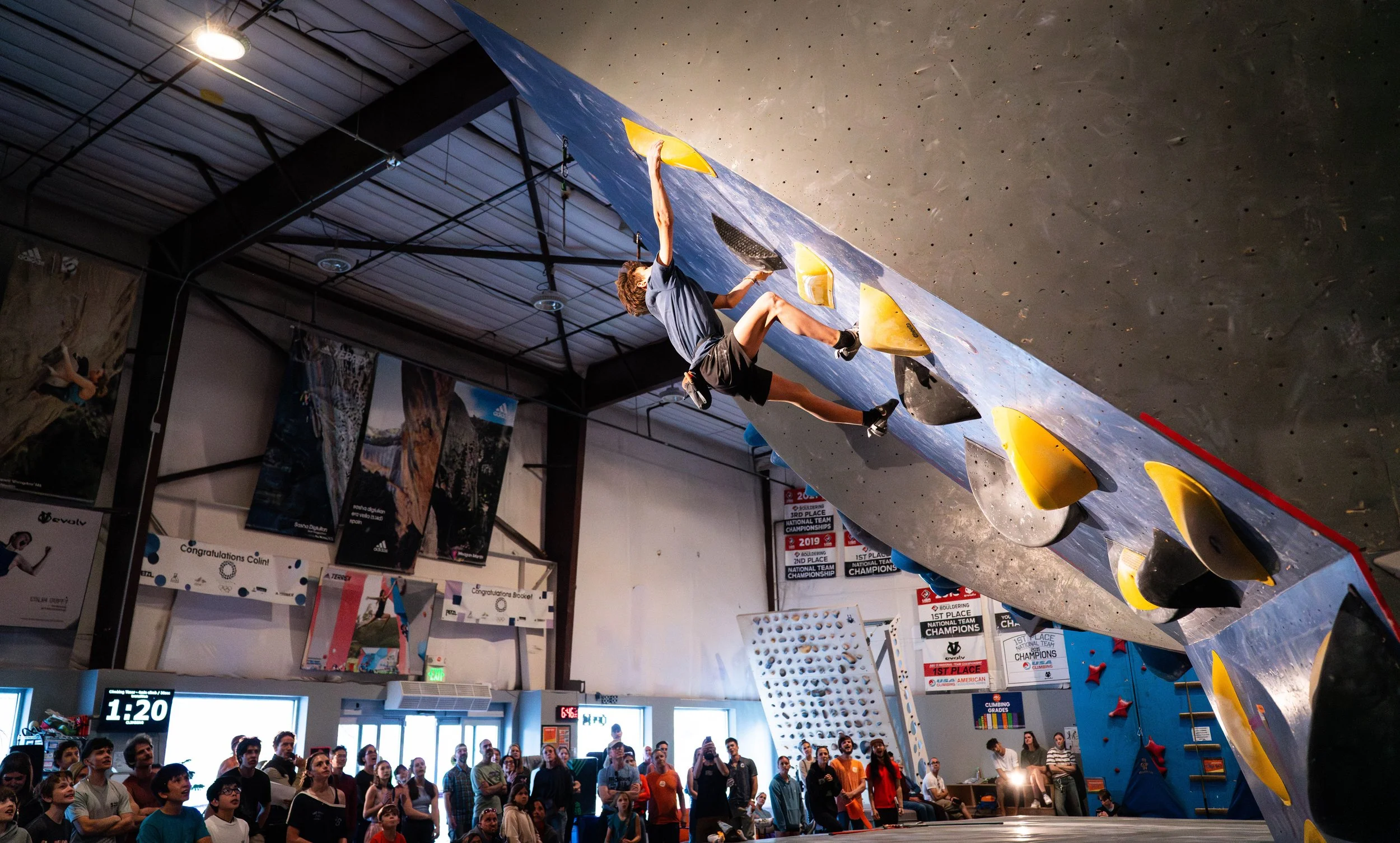 Climber on the tall overhanging wall above a crowd at ABC Kids Climbing, a 7,200 square foot youth climbing gym in Boulder, CO