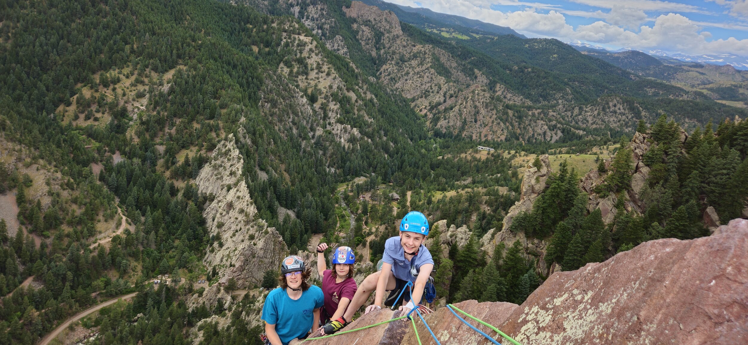 Three smiling climbers in helmets on a summit ledge high above a forested canyon during a multi-pitch climb