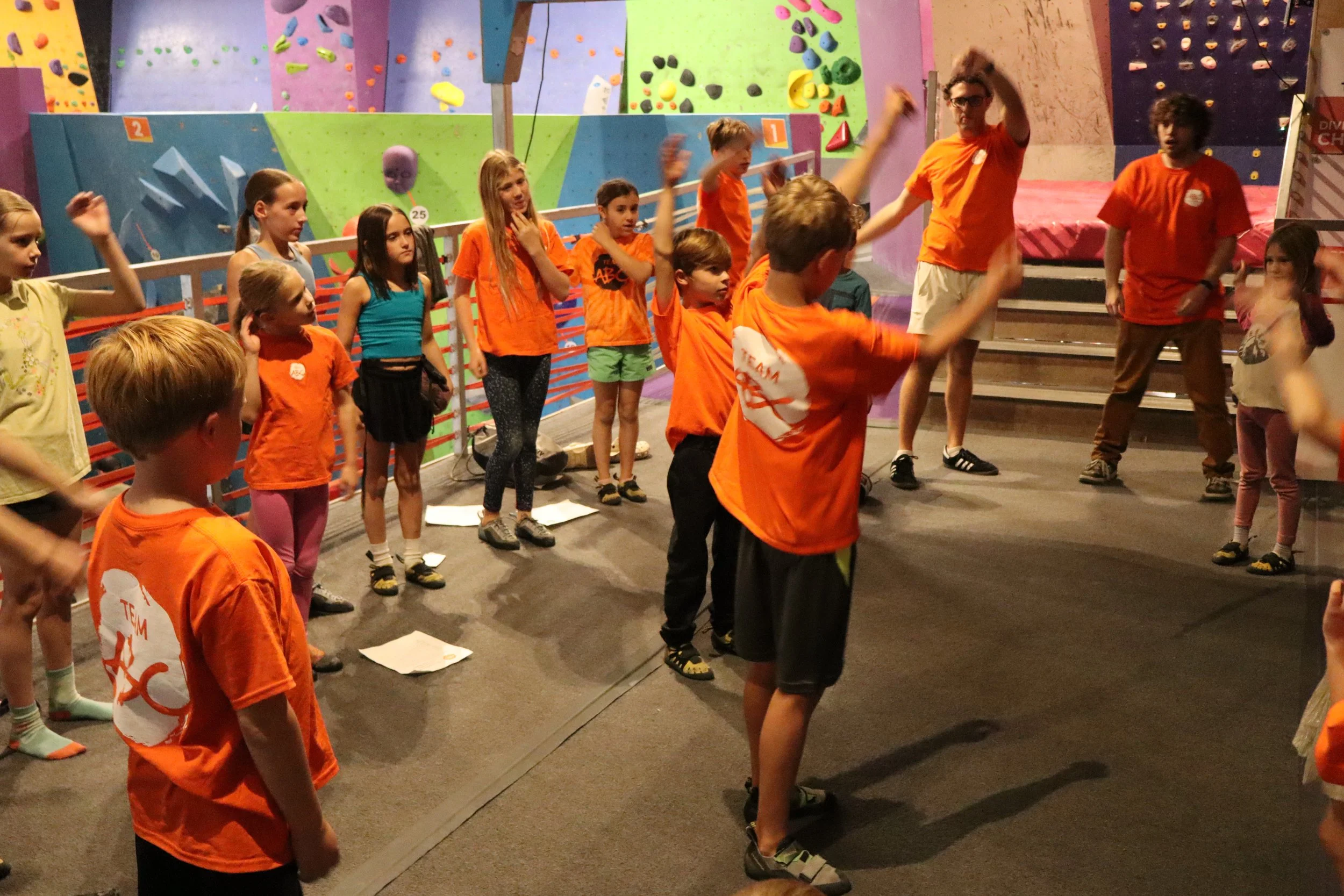 Kids in orange shirts warming up together in a circle during an ABC Kids climbing class in Boulder