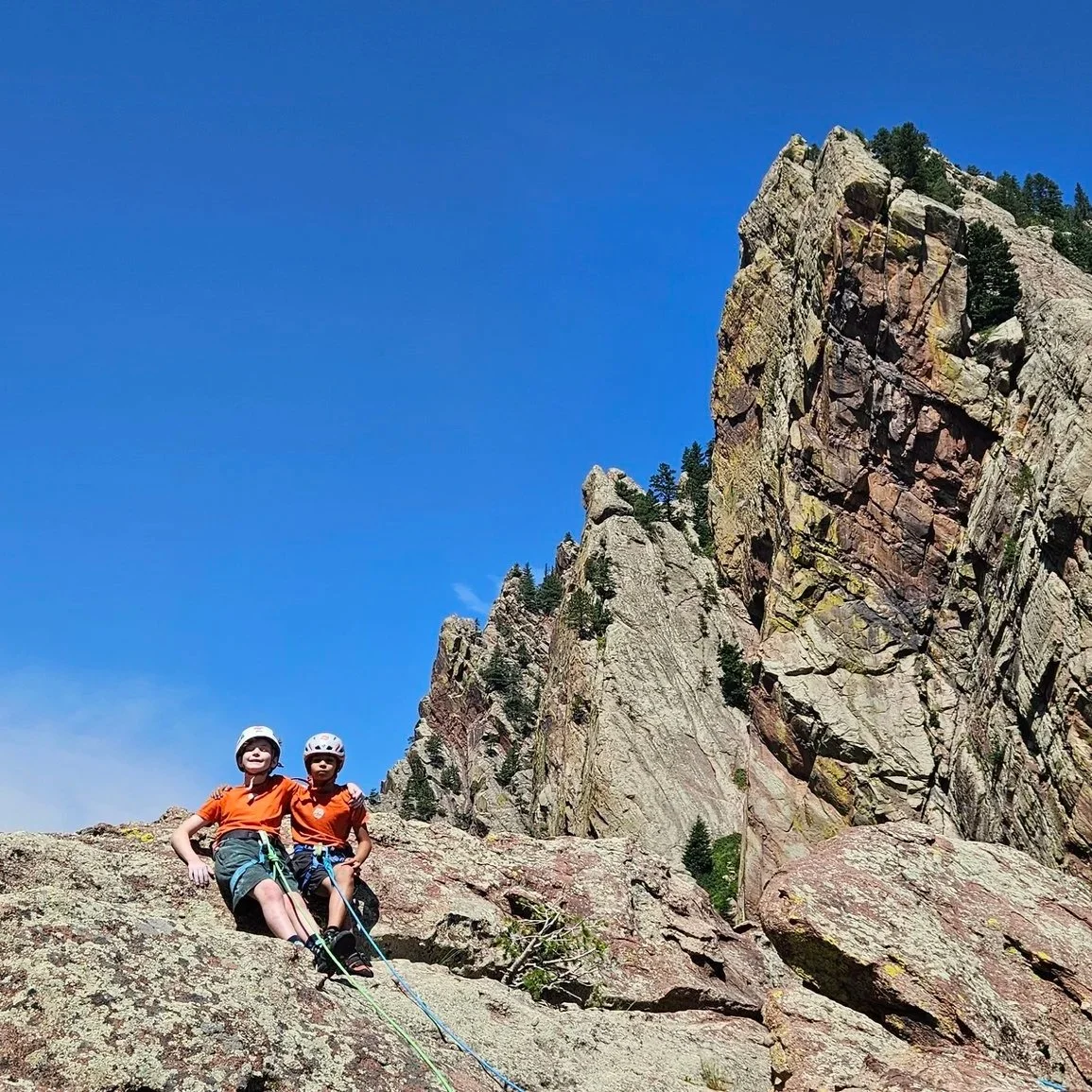 Two young climbers in helmets and harnesses below towering sandstone cliffs in Eldorado Canyon