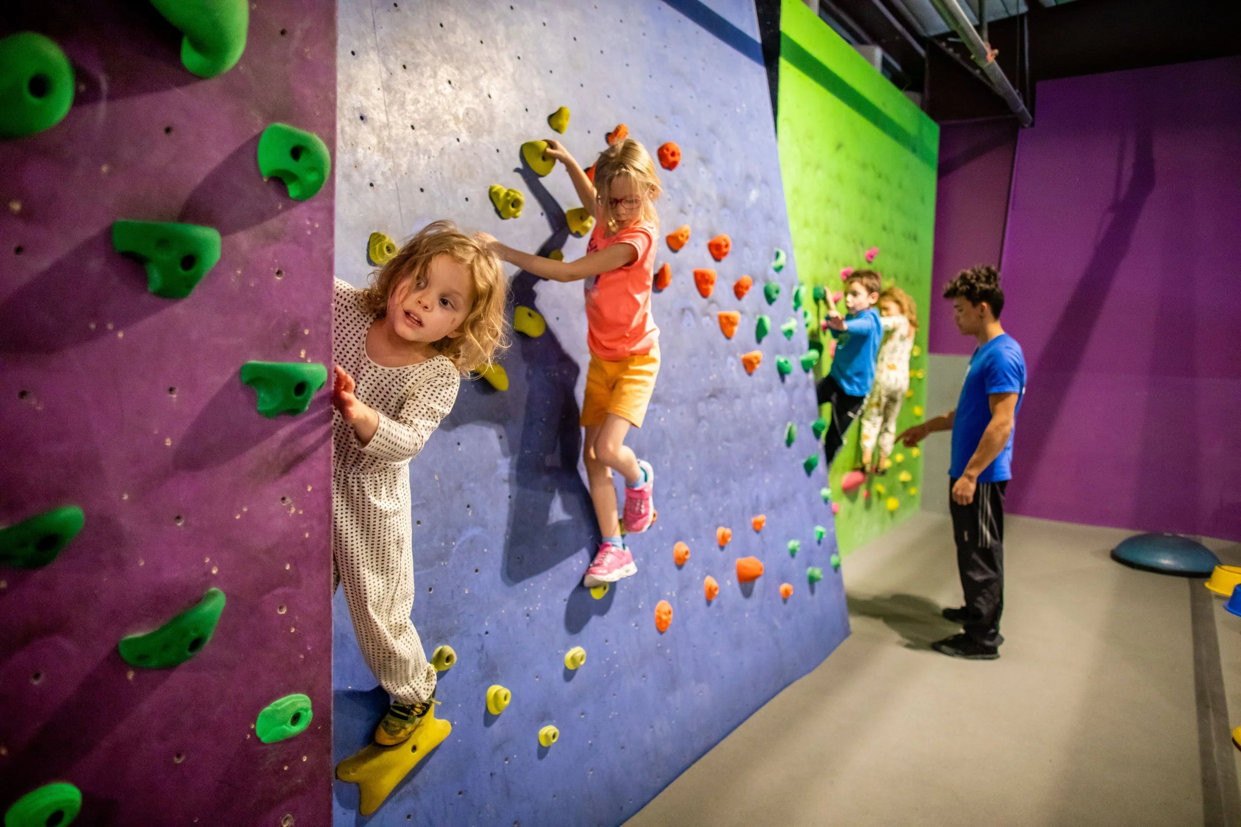 Two young girls climbing a purple kids climbing wall with green holds during a daytime class at ABC Kids Climbing