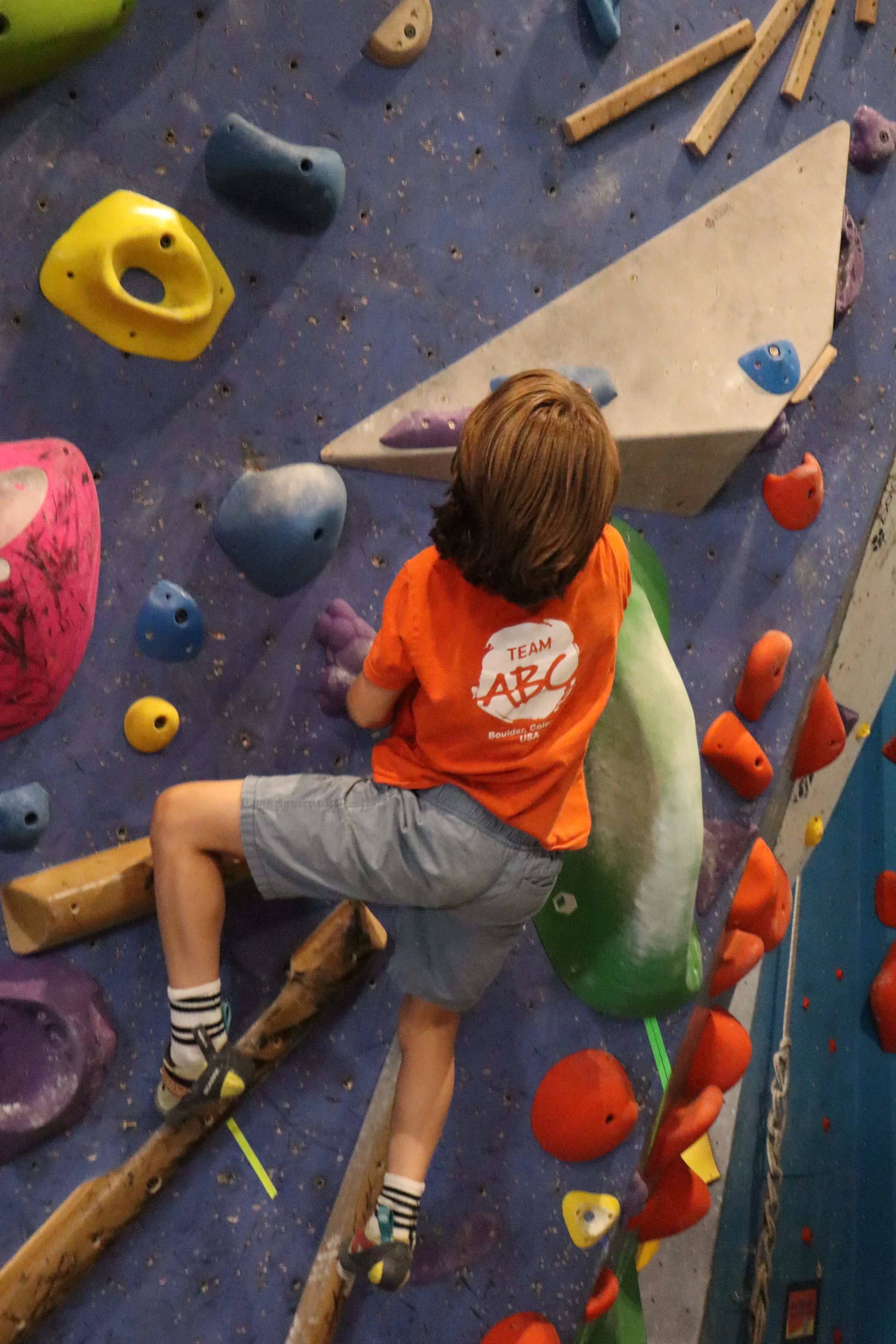 Young climber in a Team ABC shirt reaching for holds during an adaptive climbing session at ABC Kids Climbing in Boulder