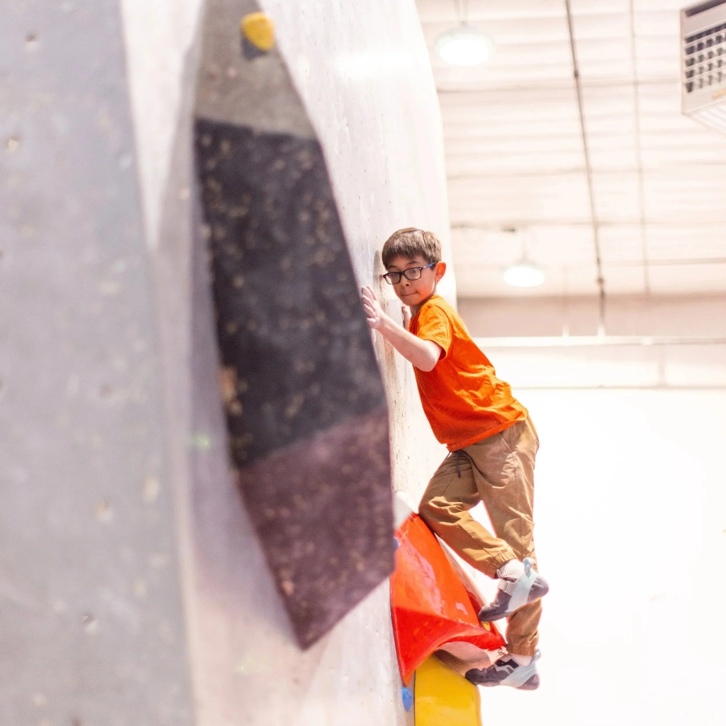 Boy with glasses in an orange shirt climbing a boulder at ABC Kids Climbing in Boulder