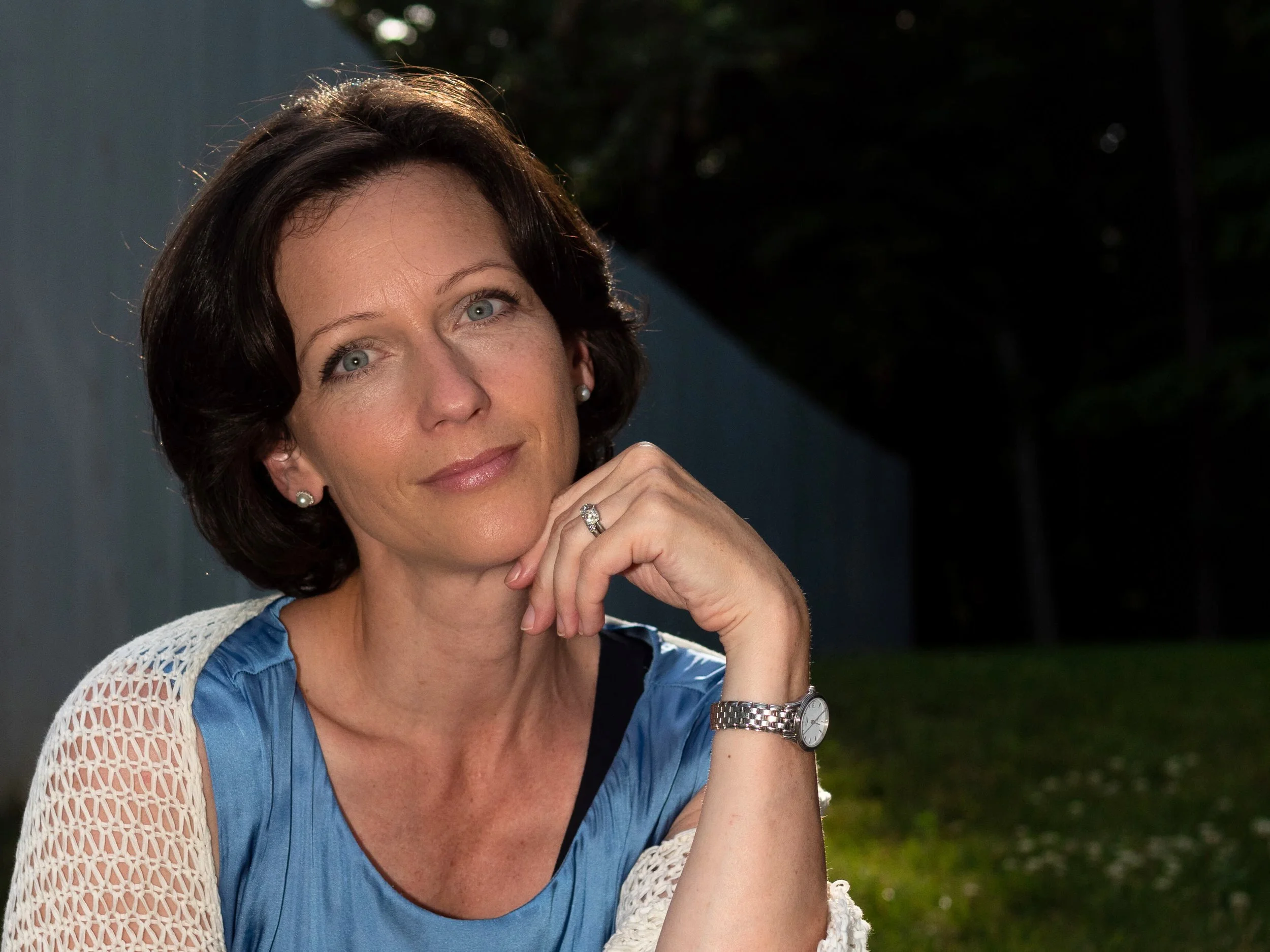 A woman with short dark hair and blue eyes, Sasha Mrak Hendrickson, wearing a blue top with a white crochet shawl, sitting outdoors at sunset, resting her chin on her hand, with a thoughtful expression.
