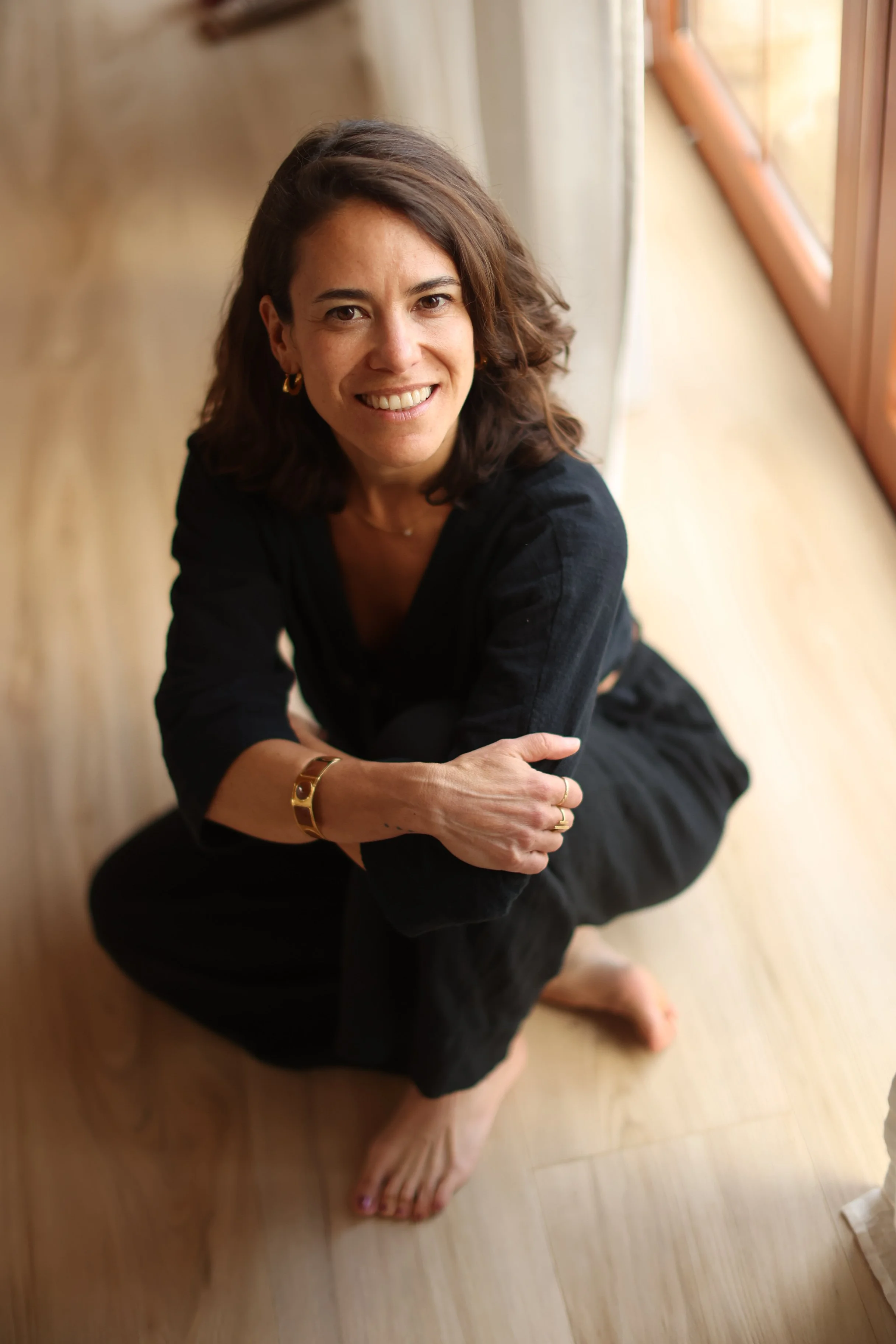 A woman with wavy brown hair, sitting on a wooden floor by a window, smiling at the camera, wearing a black long-sleeve top and gold jewelry, with her arms crossed and legs folded.