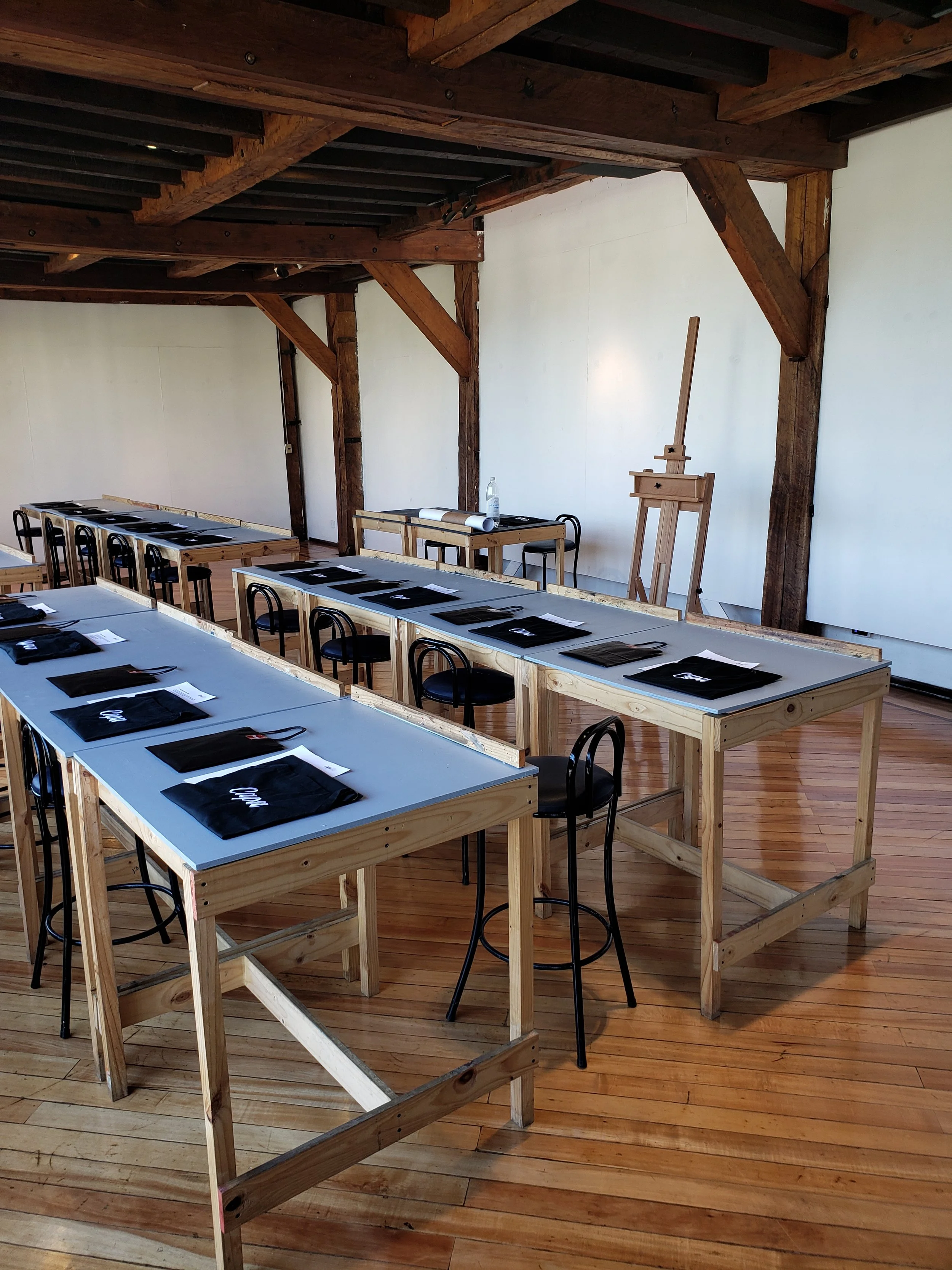 Aula de pintura con mesas de madera, sillas negras y caballetes en una sala con paredes blancas y techo de madera expuesta.