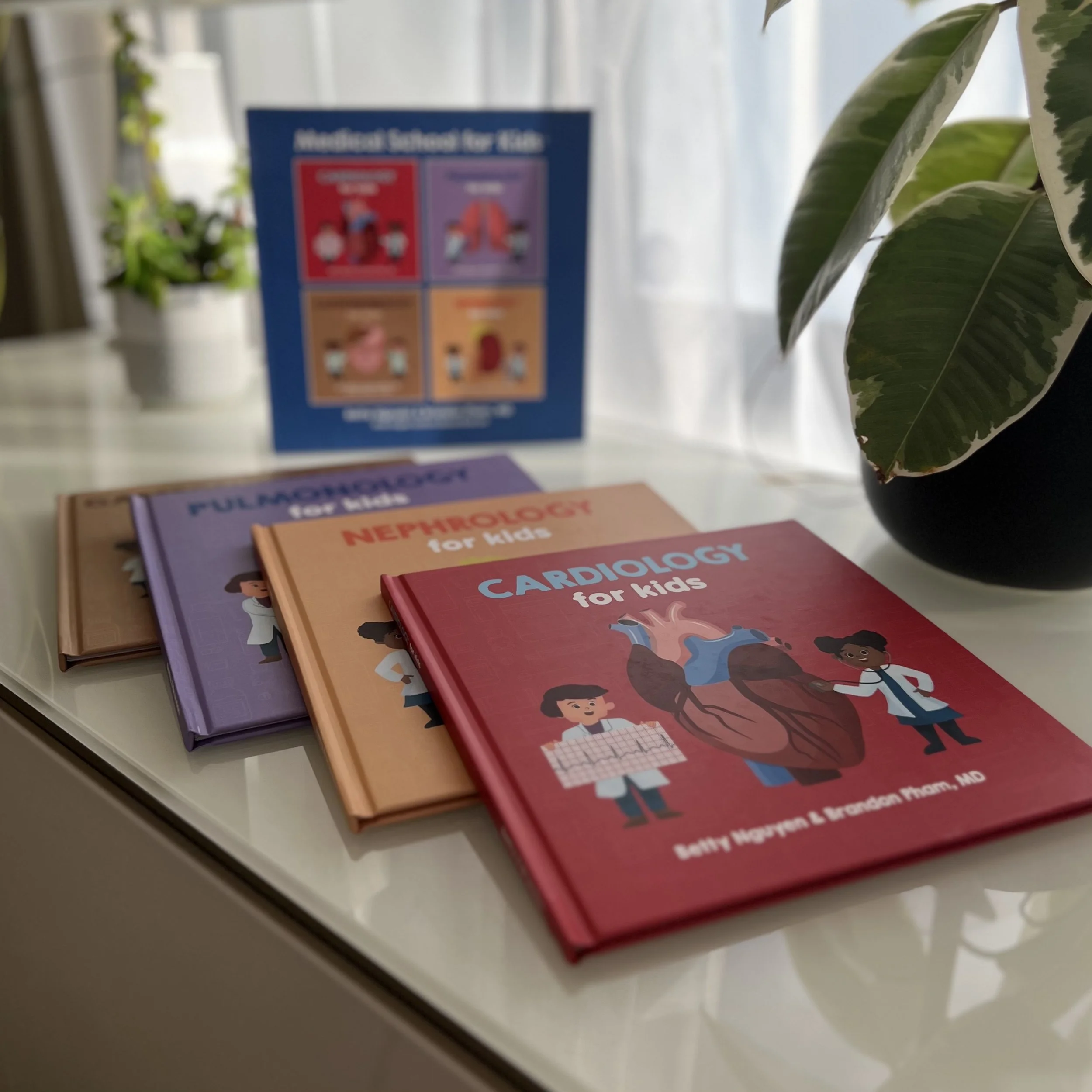 A display of colorful children's medical books on a white surface, including titles such as 'Cardiology for Kids', 'Nephrology for Kids', and 'Pulmonology for Kids', with a plant in a black pot and a blurred educational poster in the background.