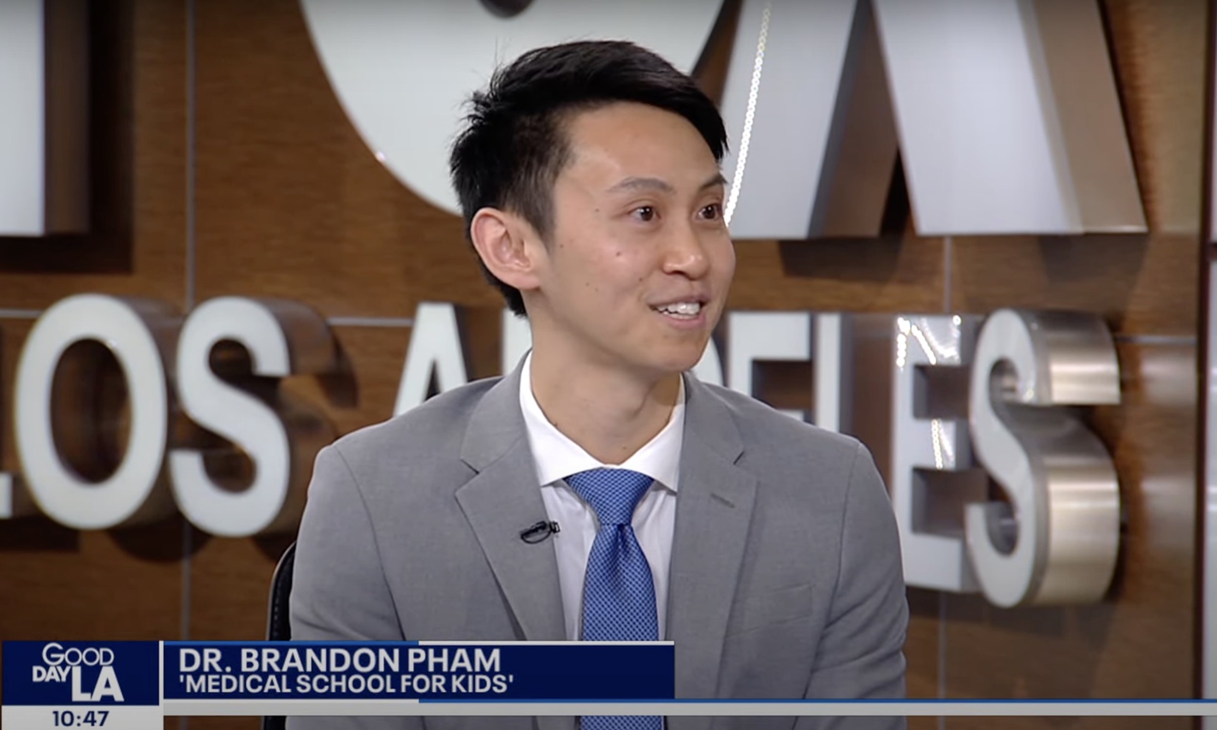 A man in a gray suit and blue tie speaking during a TV interview, with a news banner that says 'Dr. Brandon Pham, Medical School for Kids'.