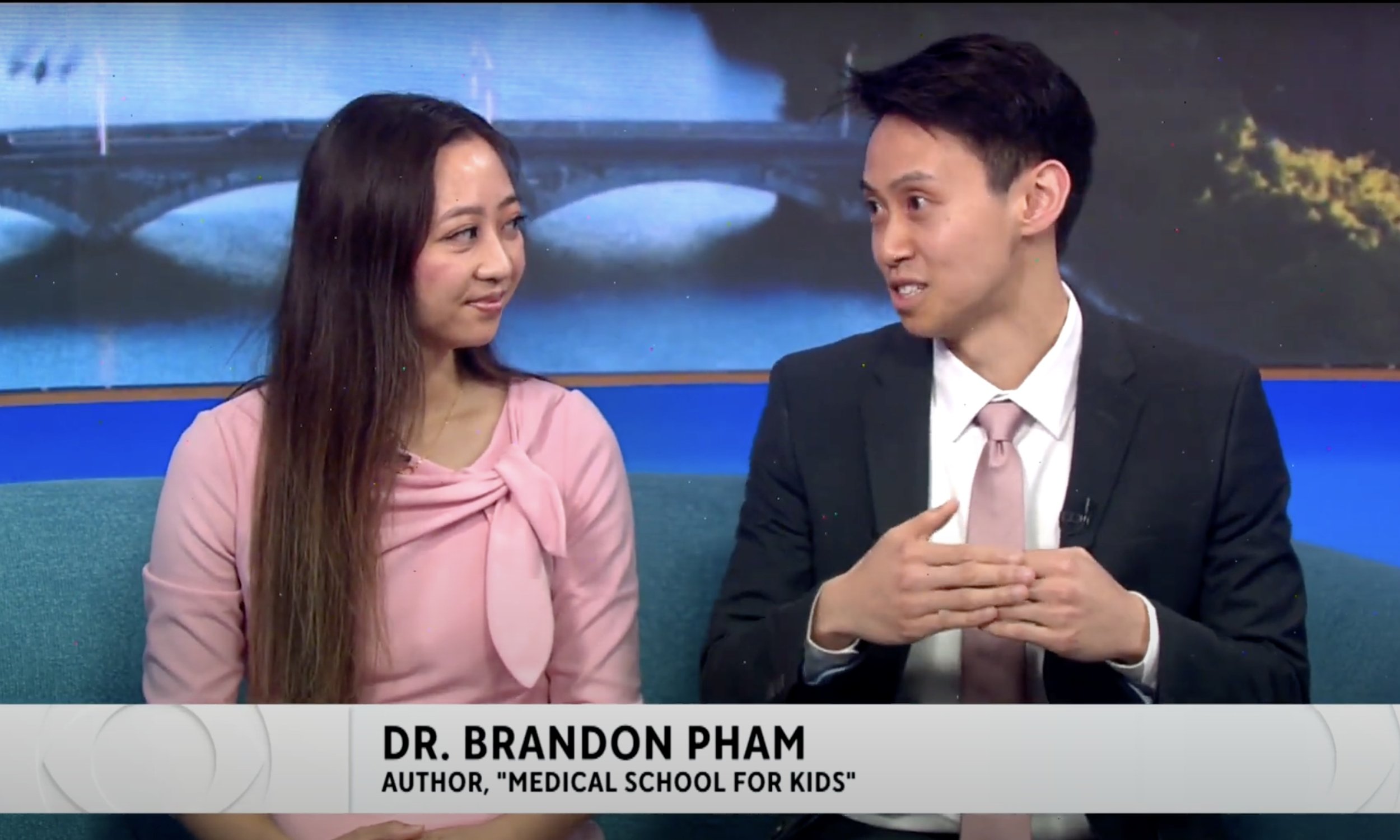 Two people, a woman in a pink dress and a man in a suit, sitting and talking on a TV set. The woman has long brown hair, and the man has short dark hair. A news banner at the bottom shows the man's name, Dr. Brandon Pham, and his title as author of "