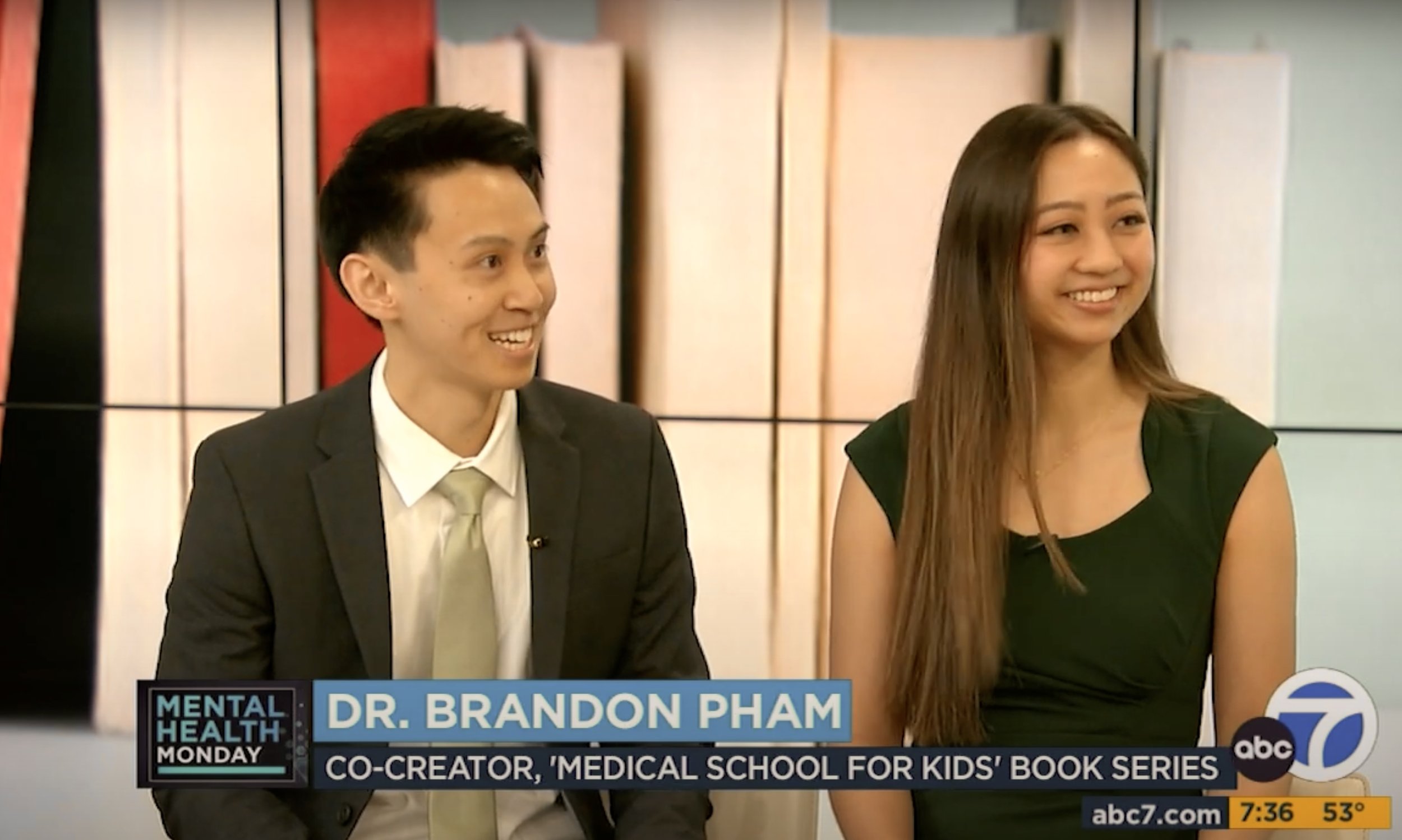 Two people, a man in a suit and a woman in a black dress, sitting and smiling during a television interview with a graphic overlay indicating the interviewee is Dr. Brandon Pham, co-creator of a children's book series about medical school, on ABC 7 N