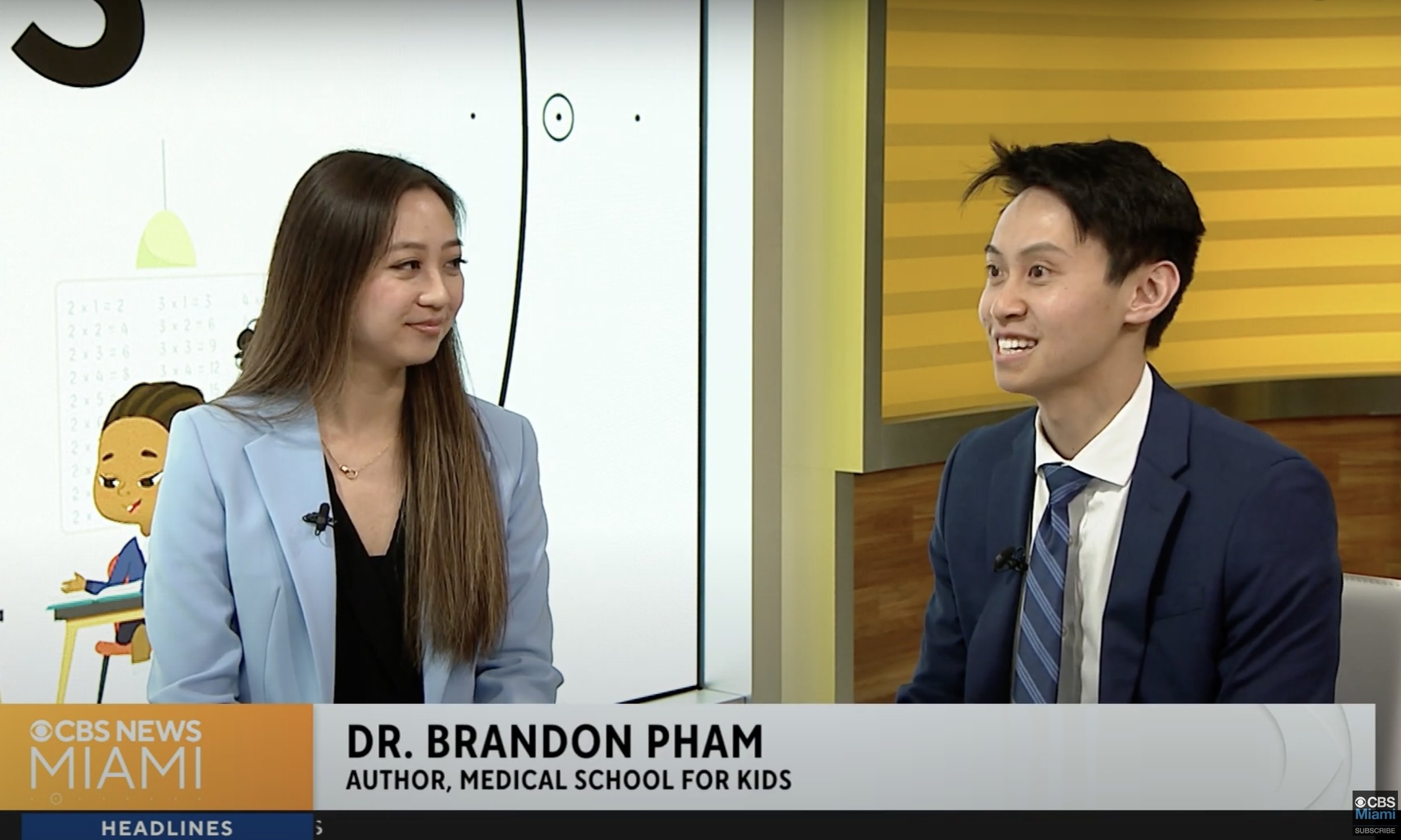 A woman and a man in business attire sitting in a television studio during an interview, with a screen displaying cartoon characters behind them. The news banner says "Dr. Brandon Pham, Co-Author of Medical School for Kids."
