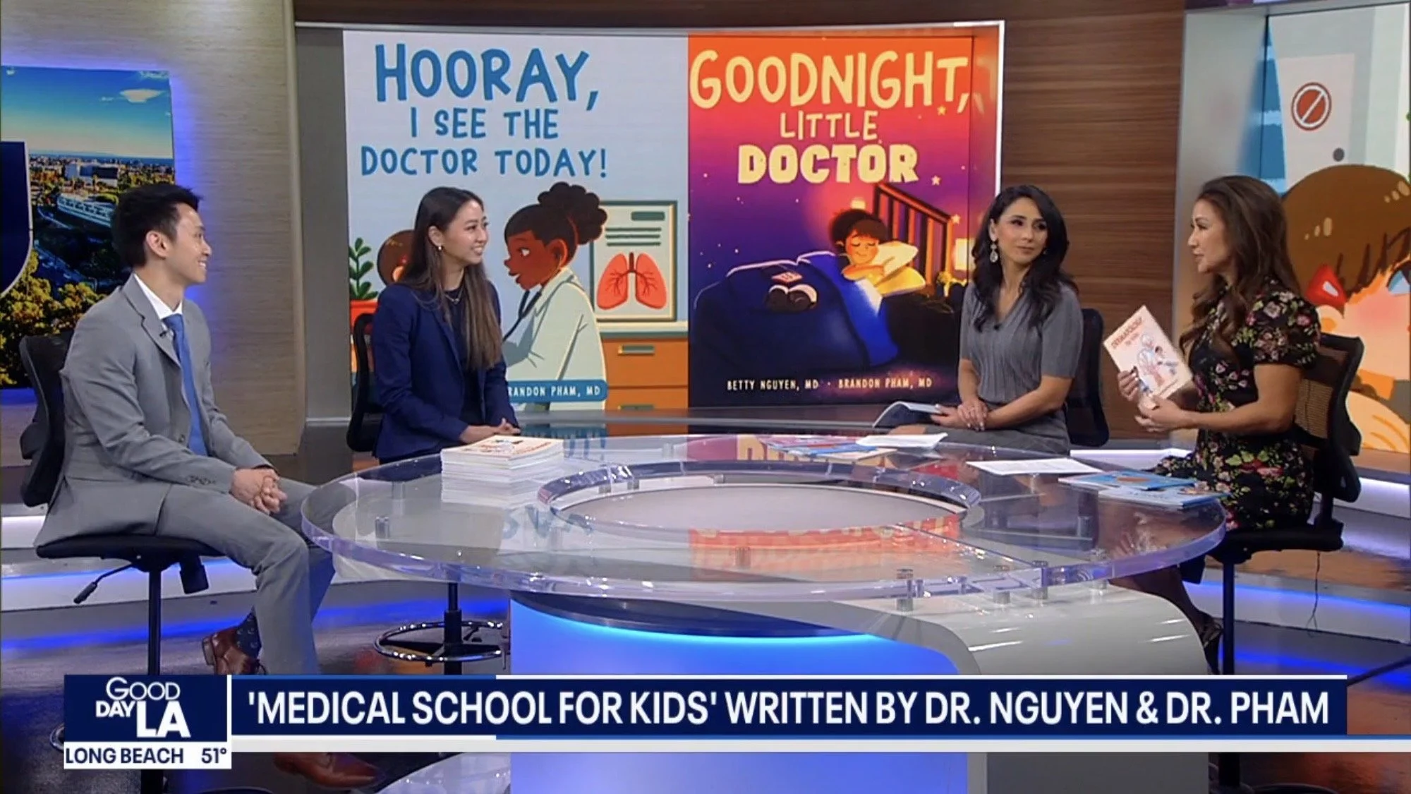 Four people seated around a glass table on a TV set, discussing a book titled 'Medical School for Kids.' Behind them are two large illustrated posters, one with a doctor and the text 'Hooray, I see the doctor today!' and the other with a child in bed and the text 'Goodnight, Little Doctor.'