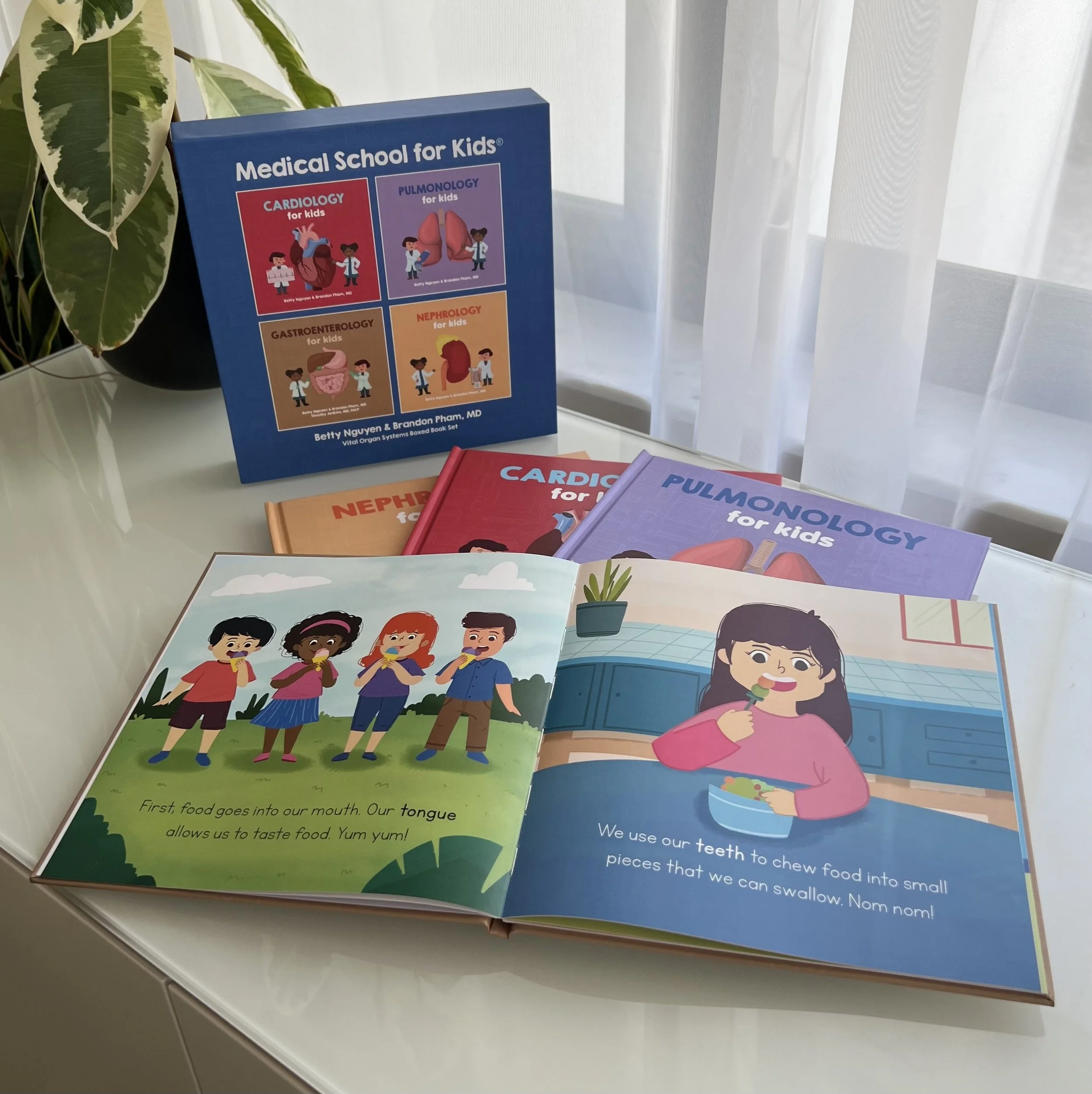 Colorful children's books on health and food open on a white table, with additional books stacked behind them, next to a potted plant with green and white leaves by a window with sheer curtains.
