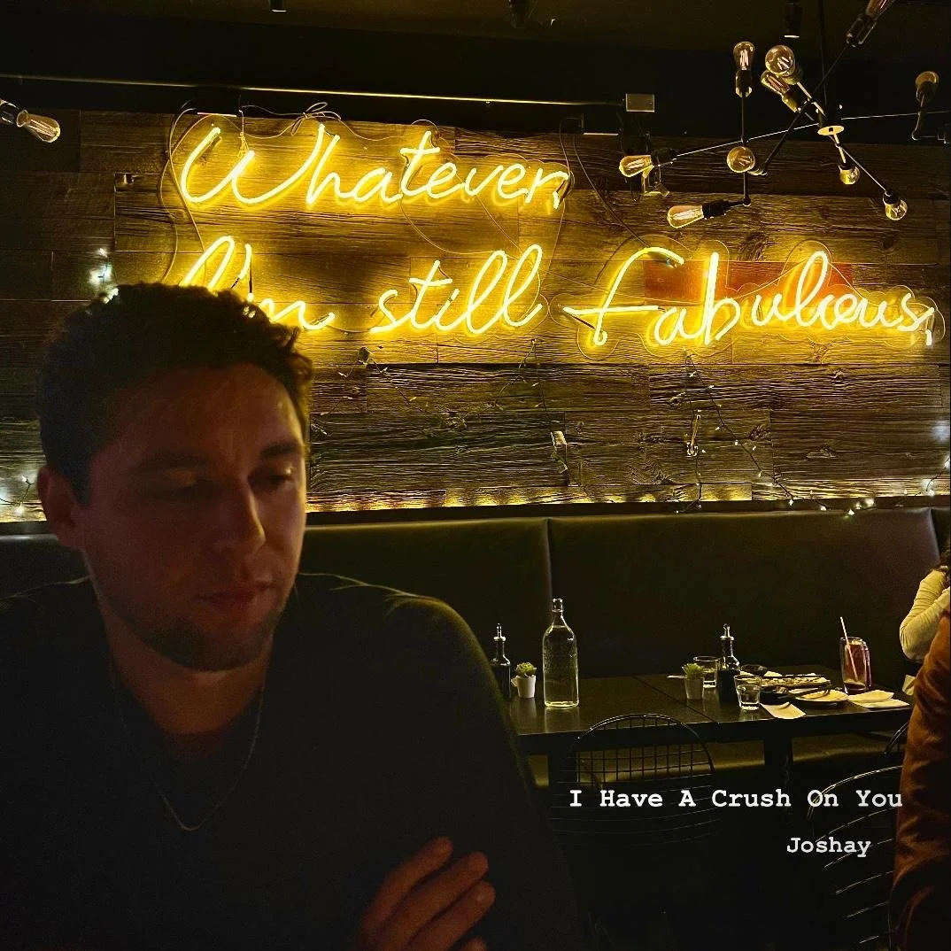 A young man (Joshay) sitting in a dimly lit restaurant with a neon sign on the wooden wall behind him that reads "Whatever You Still Fabulous." There are tables with bottles, glasses, and utensils in the background.