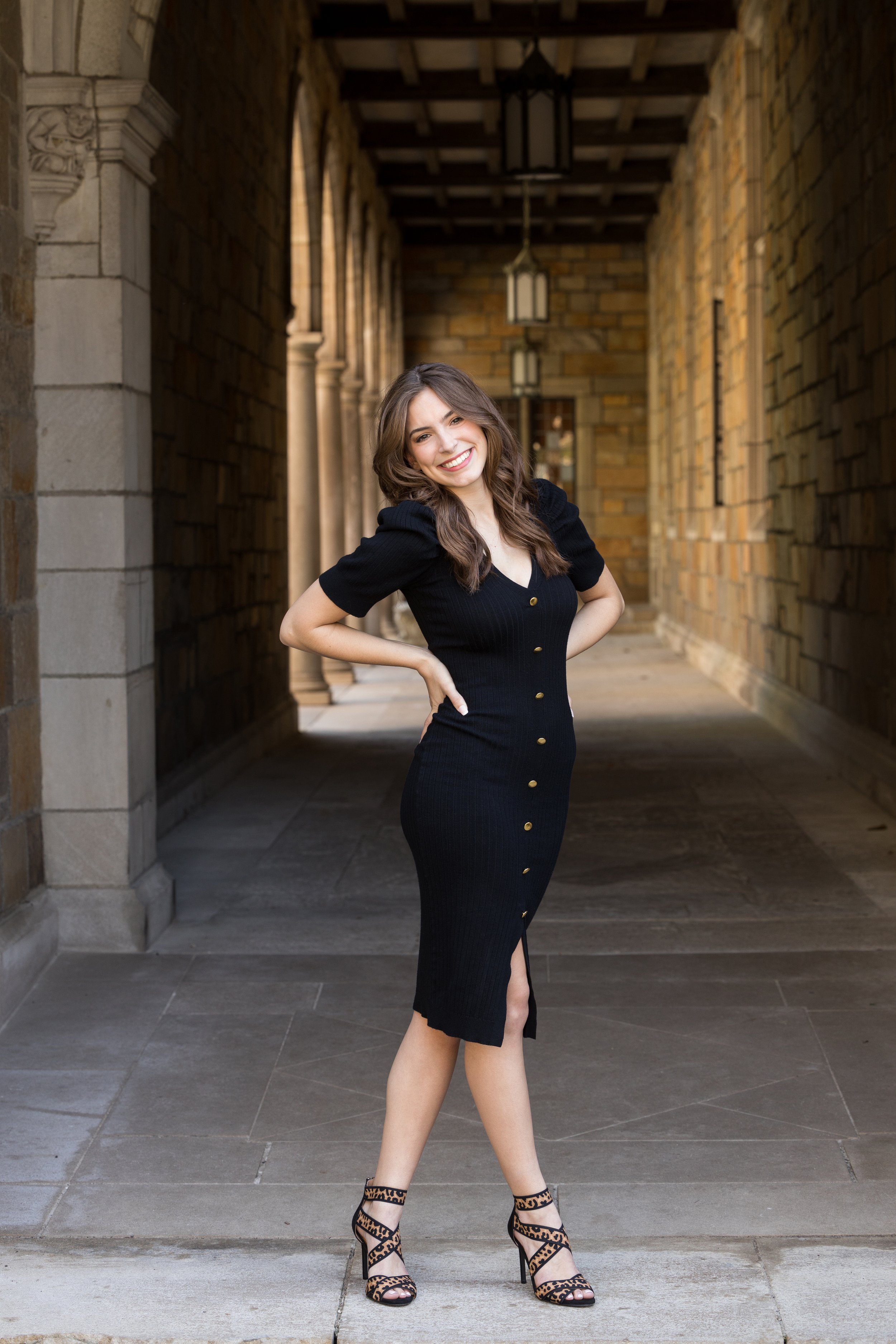 A young woman in a black dress with buttons and a slit, wearing high-heeled leopard print shoes, standing in an arched brick walkway with hanging lanterns, smiling.