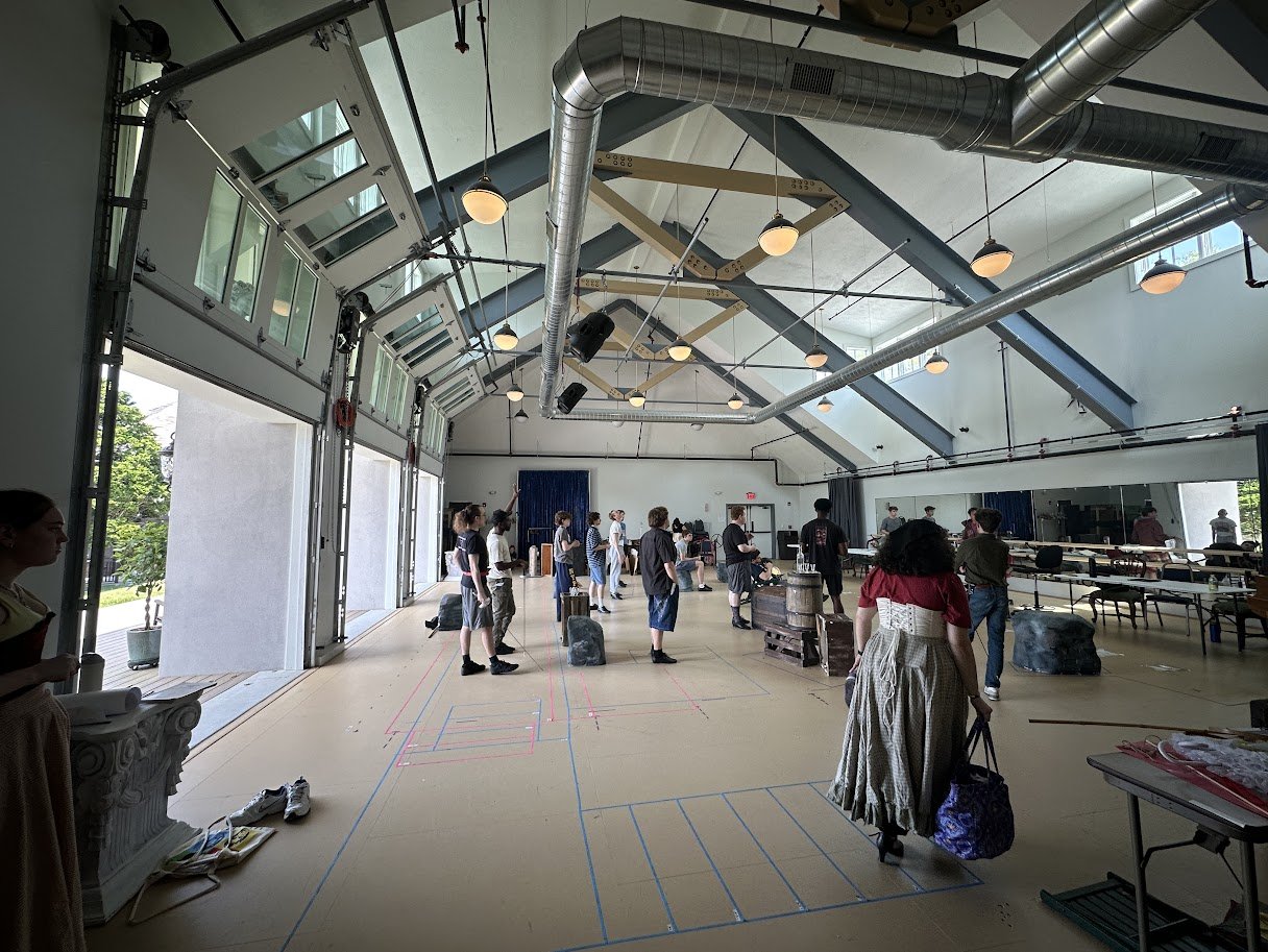 A group of people practicing yoga in a spacious studio with high ceilings, large windows, and industrial ductwork, some standing on yoga mats and others holding poses, with various props and equipment on the sides.