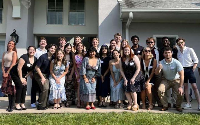Group of young adults standing outside a house, posing for a photo, with some making thumbs up and smiling.