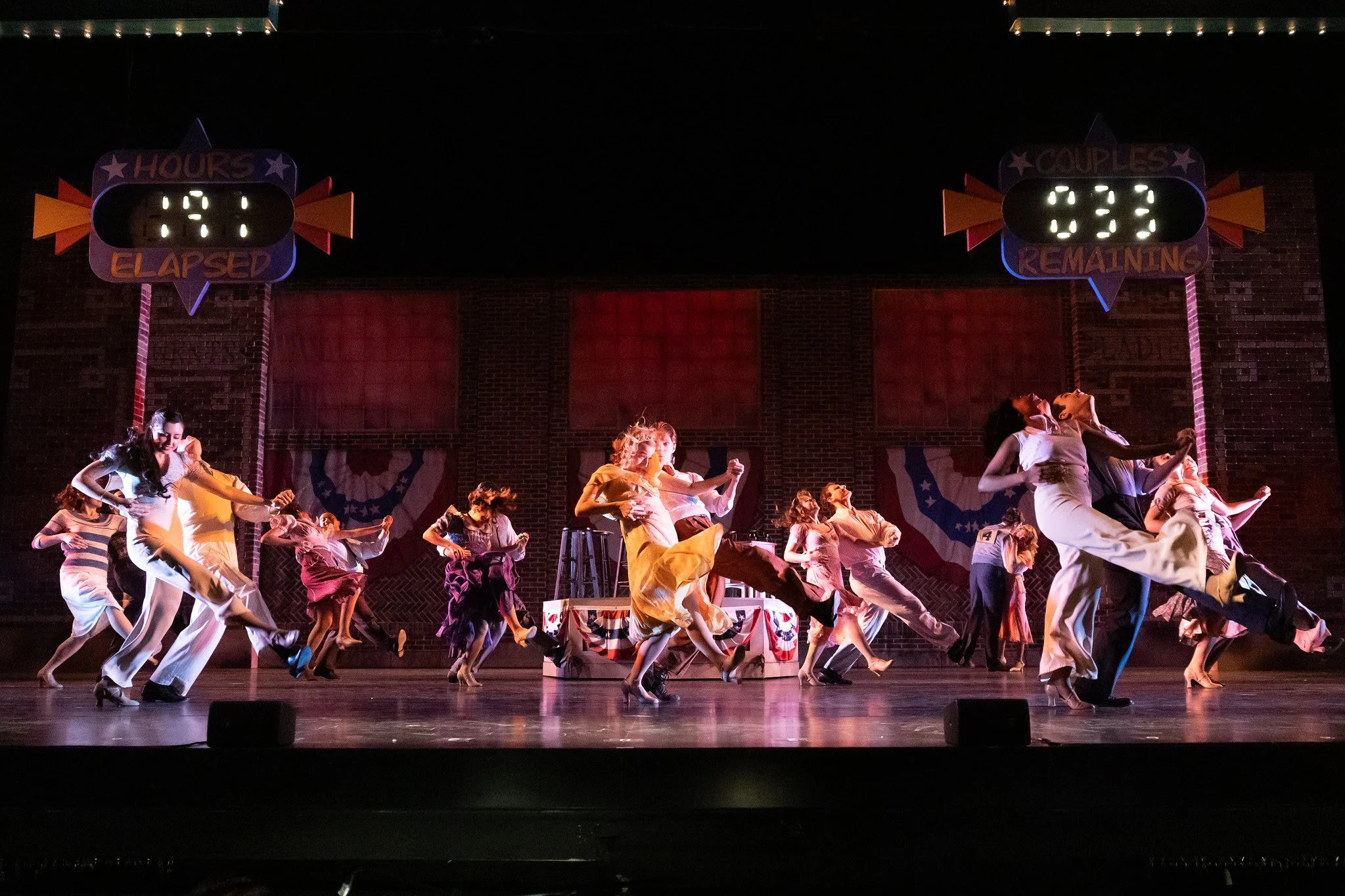 A group of dancers performing on stage in vintage costumes, with patriotic decorations and scoreboard displays indicating time elapsed and remaining.