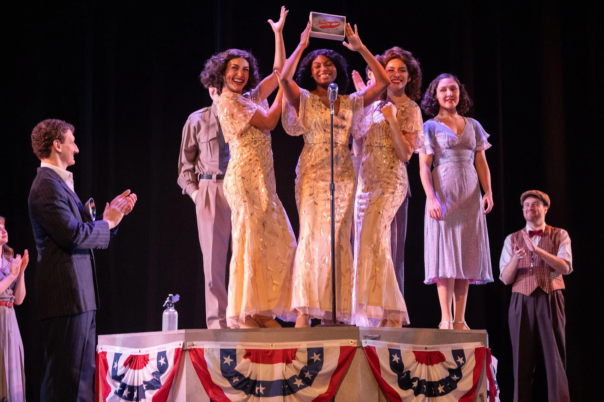 Group of people on stage celebrating, with one woman holding a box above her head, dressed in vintage clothing, and American patriotic decorations.