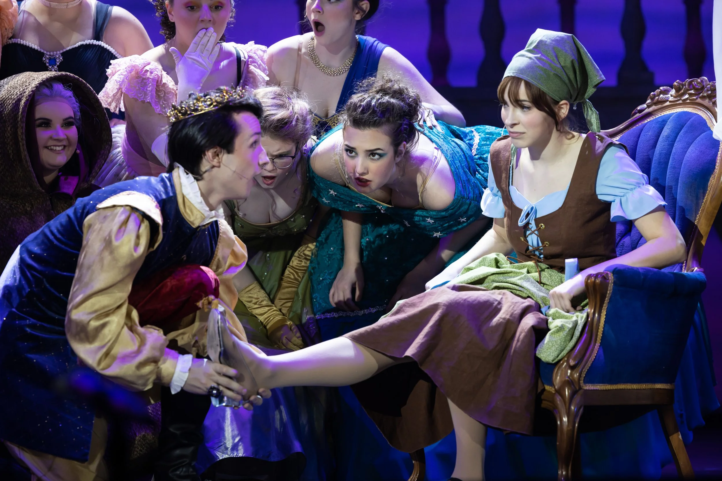 The scene from a theatrical performance, with actors dressed in medieval or fairy-tale costumes, gathered around a woman seated in a vintage chair. The woman appears to be injured or in distress, while the actors show concern and curiosity.