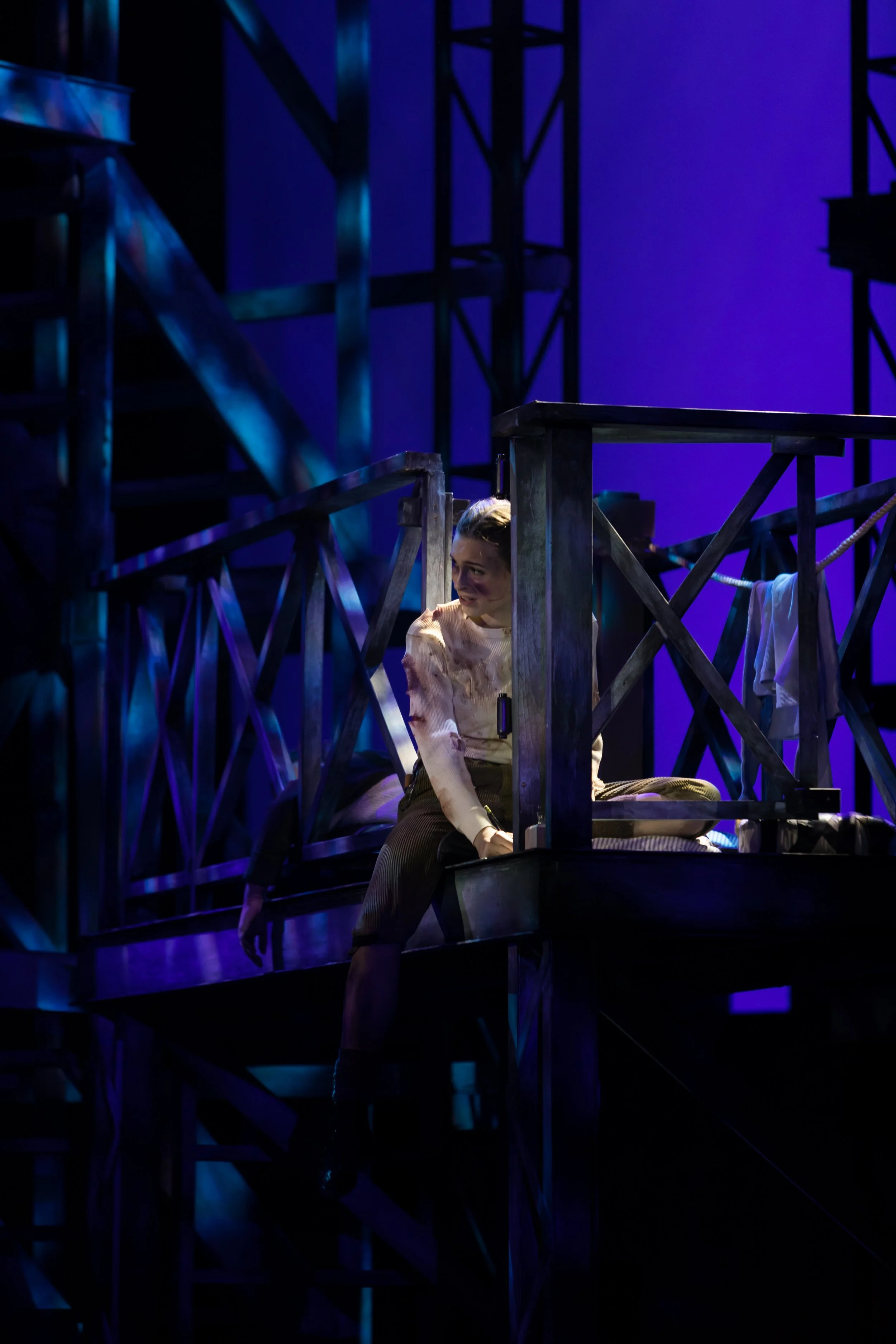 A stage scene with a performer sitting on a wooden platform, illuminated by dramatic lighting with purple and blue hues. The performer is wearing a light-colored blouse and dark pants, with a mischievous or playful expression.