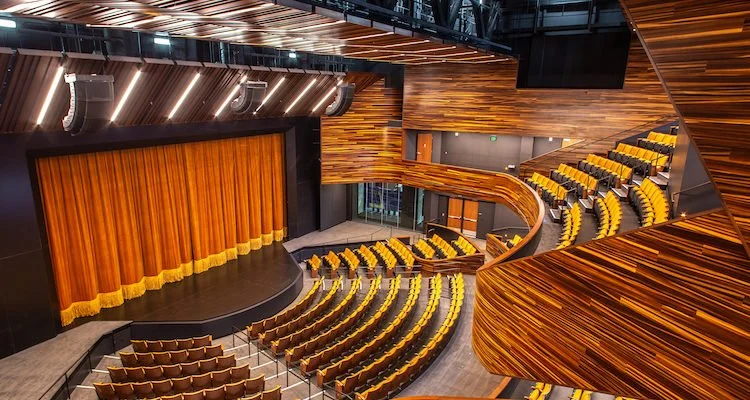 Interior of a modern theater or auditorium with wooden paneling, yellow seats, and a stage with a closed curtain.