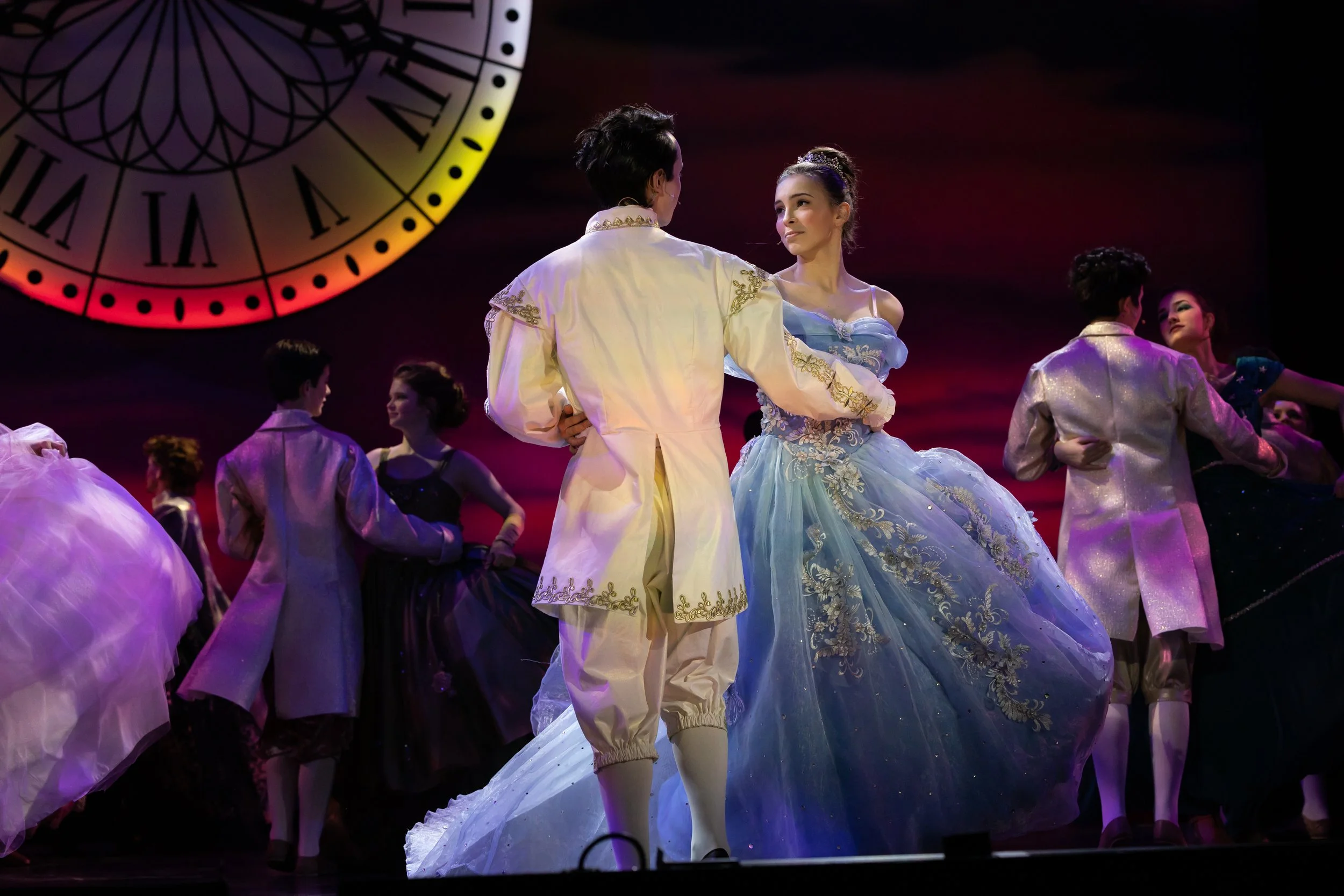 A stage scene with several performers in elegant costumes, including a woman in a light blue gown and a man in a matching cream outfit, dancing while other dancers watch or dance around them. The background features a large clock with Roman numerals.