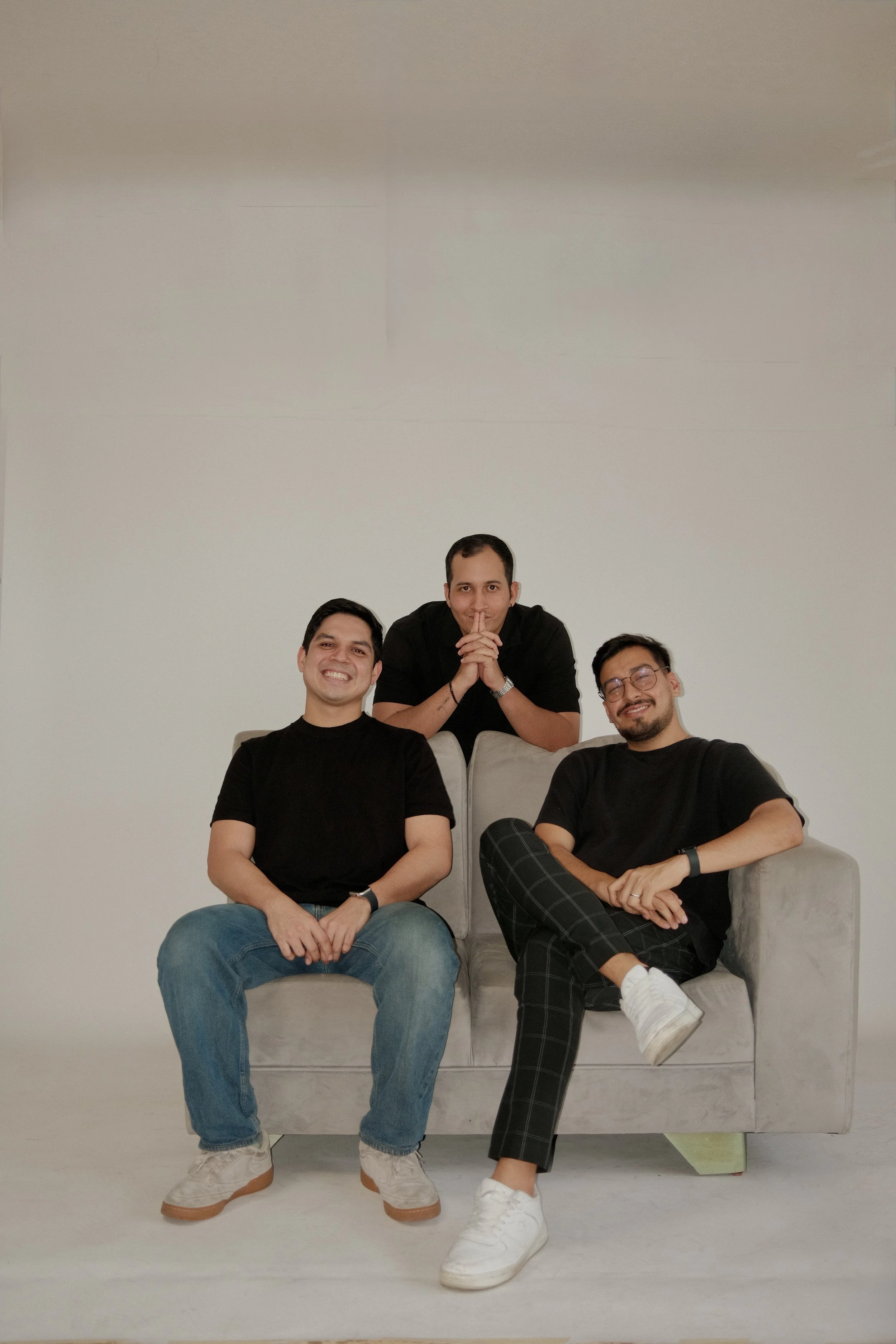 Tres hombres sentados y de pie en un sillón en un fondo blanco. Todos llevan ropa negra y sonriendo a la cámara.