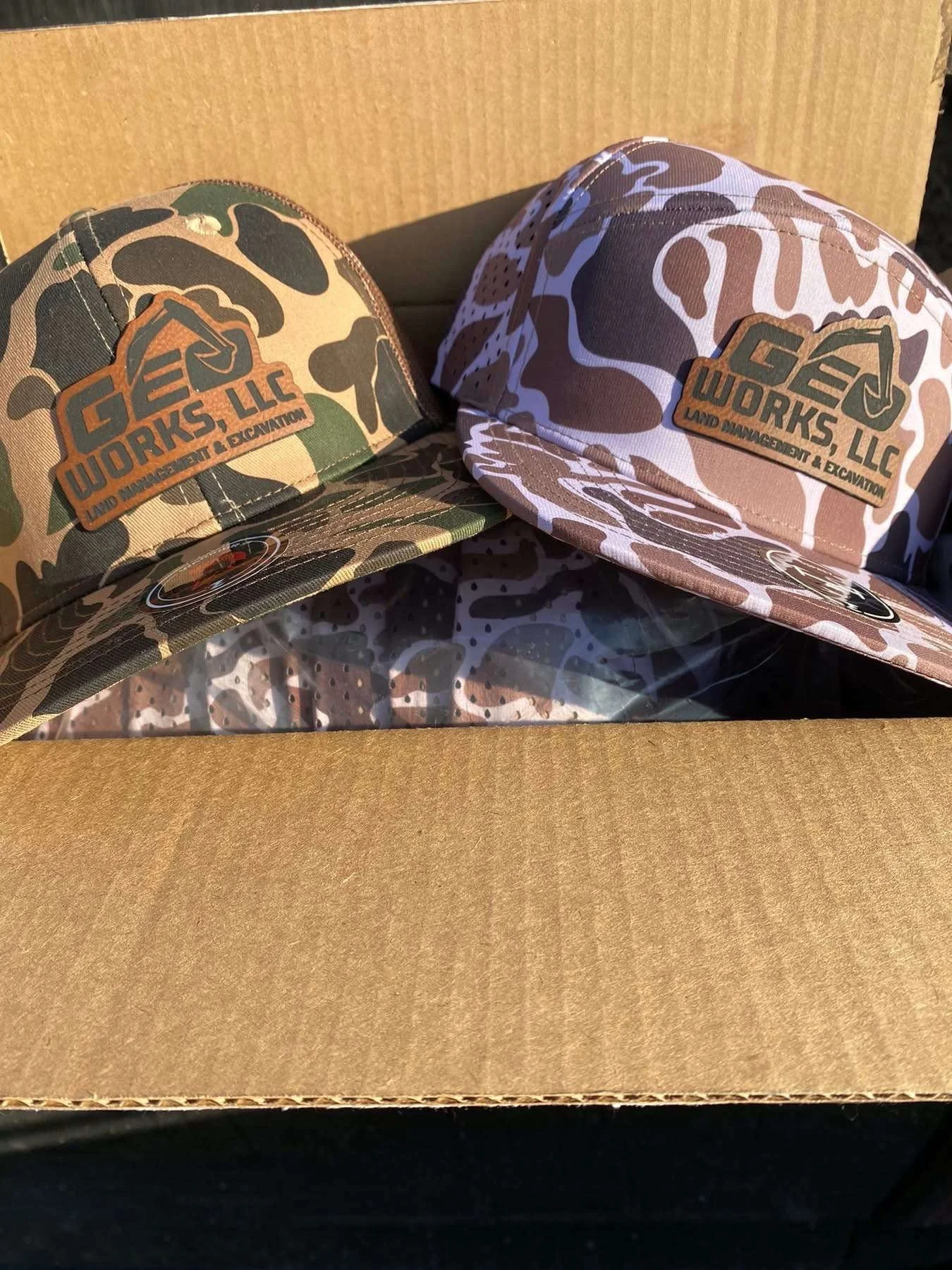 Two camouflage-patterned caps with leather patches reading 'GEOWORKS, LLC LAND MANAGEMENT & EXCAVATION' 