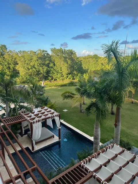 A view of resort grounds with a room below. The room has a day bed and private pool overlooking lush green grass and palm trees.