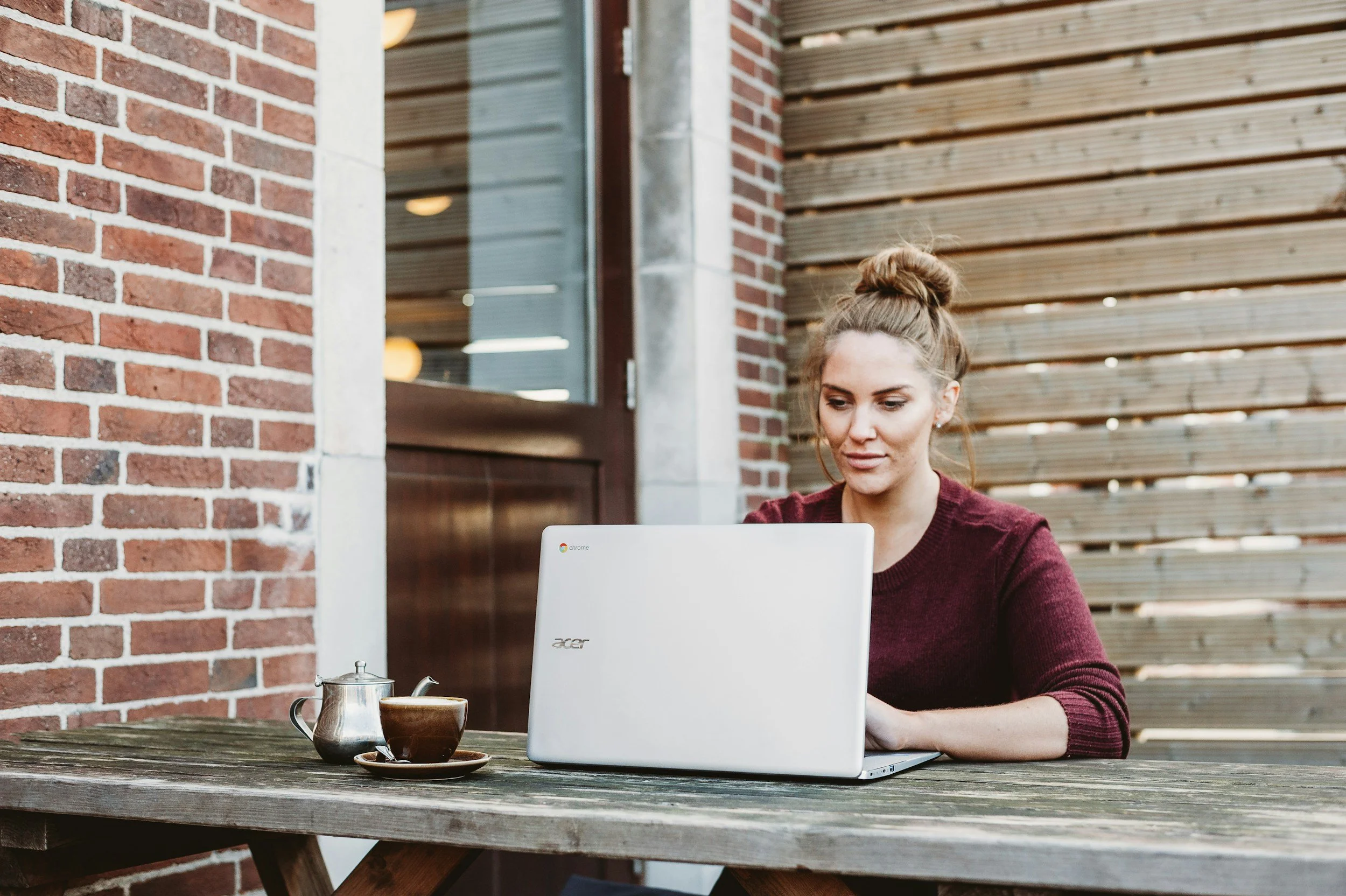 An early 30s Woman with a maroon shirt and her hair in a bun is sitting outside at a wooden table with a cup of tea. She is attending her secure, virtual therapy session working on healing her childhood trauma.