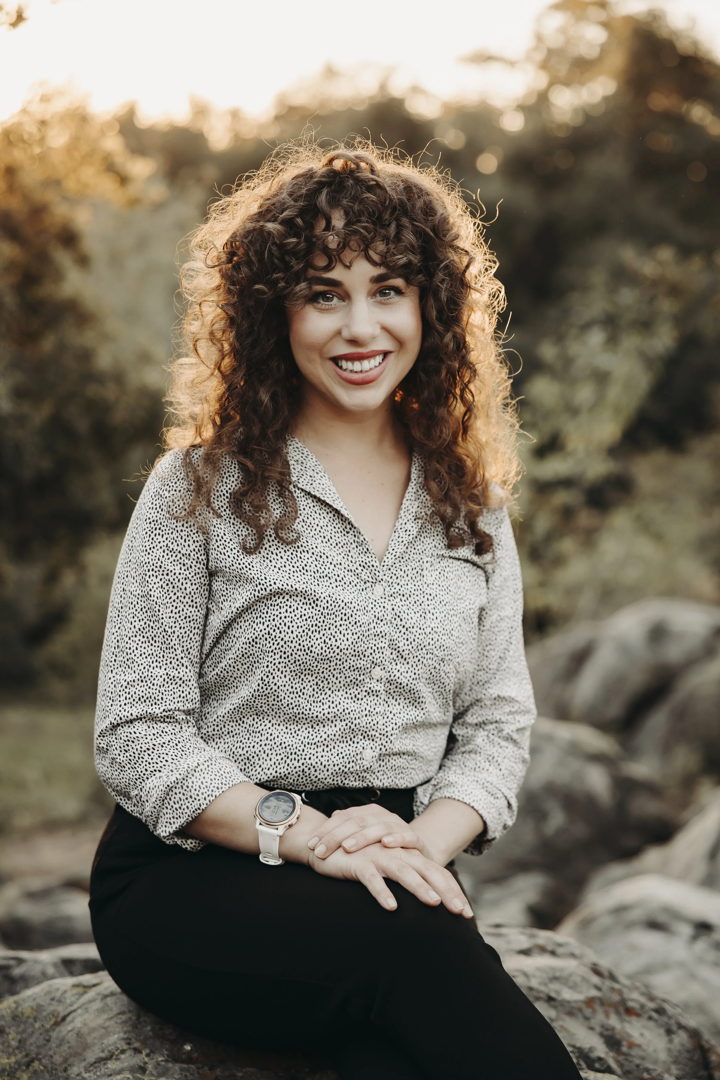 Alexa Hall is an Anxiety Therapist San Diego and is shown seated, wearing black pants, a dotted black and white shirt, she has curly hair and is smiling at the camera to welcome clients to her Anxiety Therapy practice