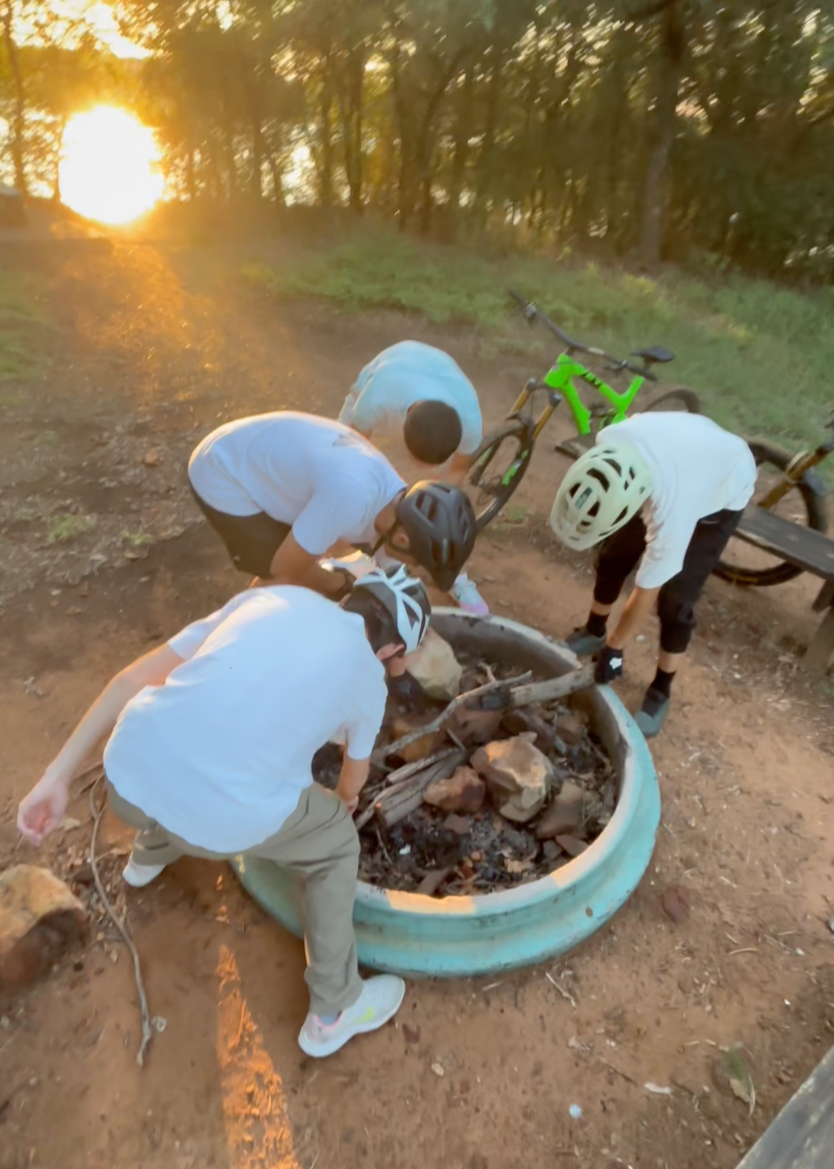 Four children wearing helmets gather around a small fire pit with rocks and sticks, tending to it in a wooded area during sunset.