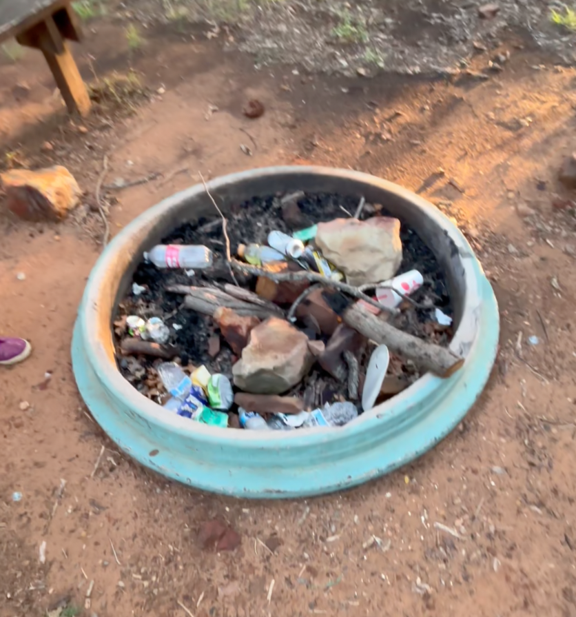 A fire pit filled with ashes, rocks, and trash including plastic bottles and cans, in an outdoor park area.