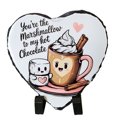 A heart-shaped chocolate candy with a cartoon illustration of a cup of hot chocolate with whipped cream, a marshmallow, and a cinnamon stick. The text reads, "You're the marshmallow to my hot chocolate," with a pink heart and a smiling marshmallow.