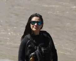 Woman wearing sunglasses and a wetsuit on a beach or shoreline.