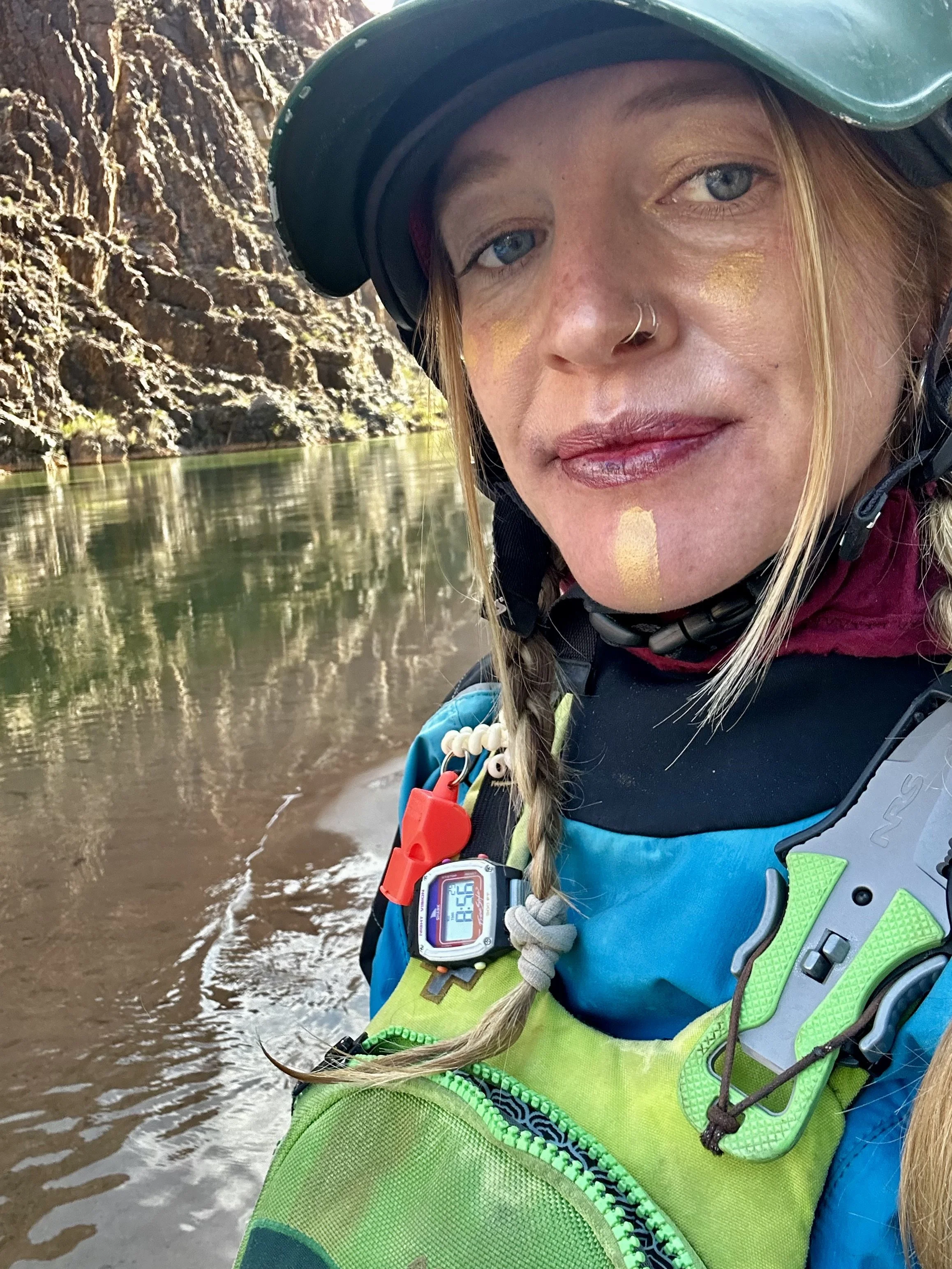 A woman in outdoor gear taking a selfie near a river with rocky cliffs in the background.