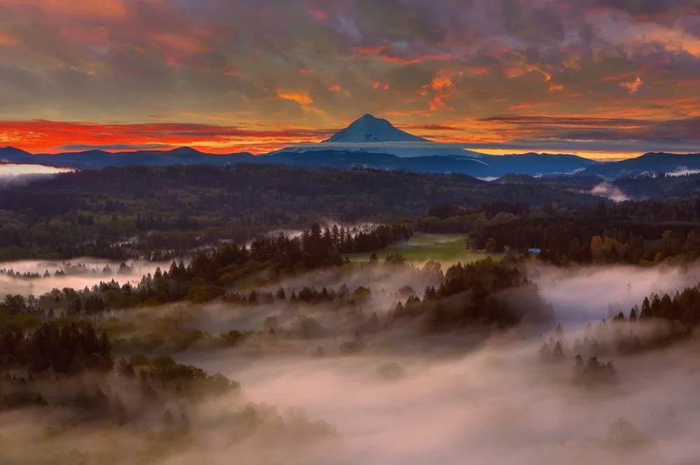 Sunrise over a mountain landscape with fog-filled valleys and a snow-capped peak in the distance.