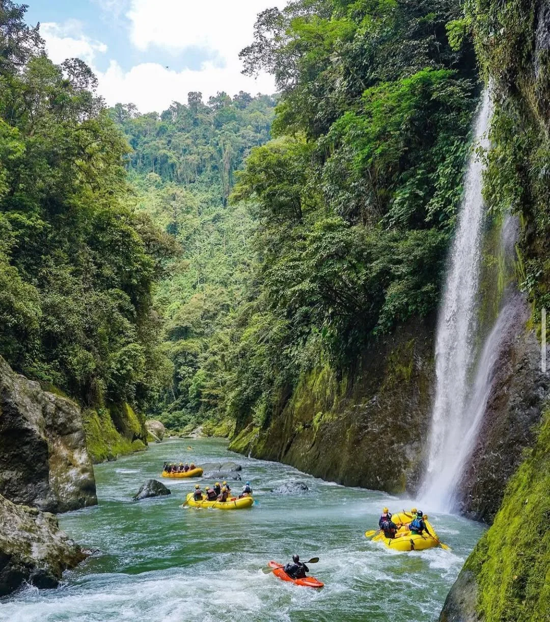 A group of people white water rafting in a river surrounded by dense green forest with a waterfall on the right side.