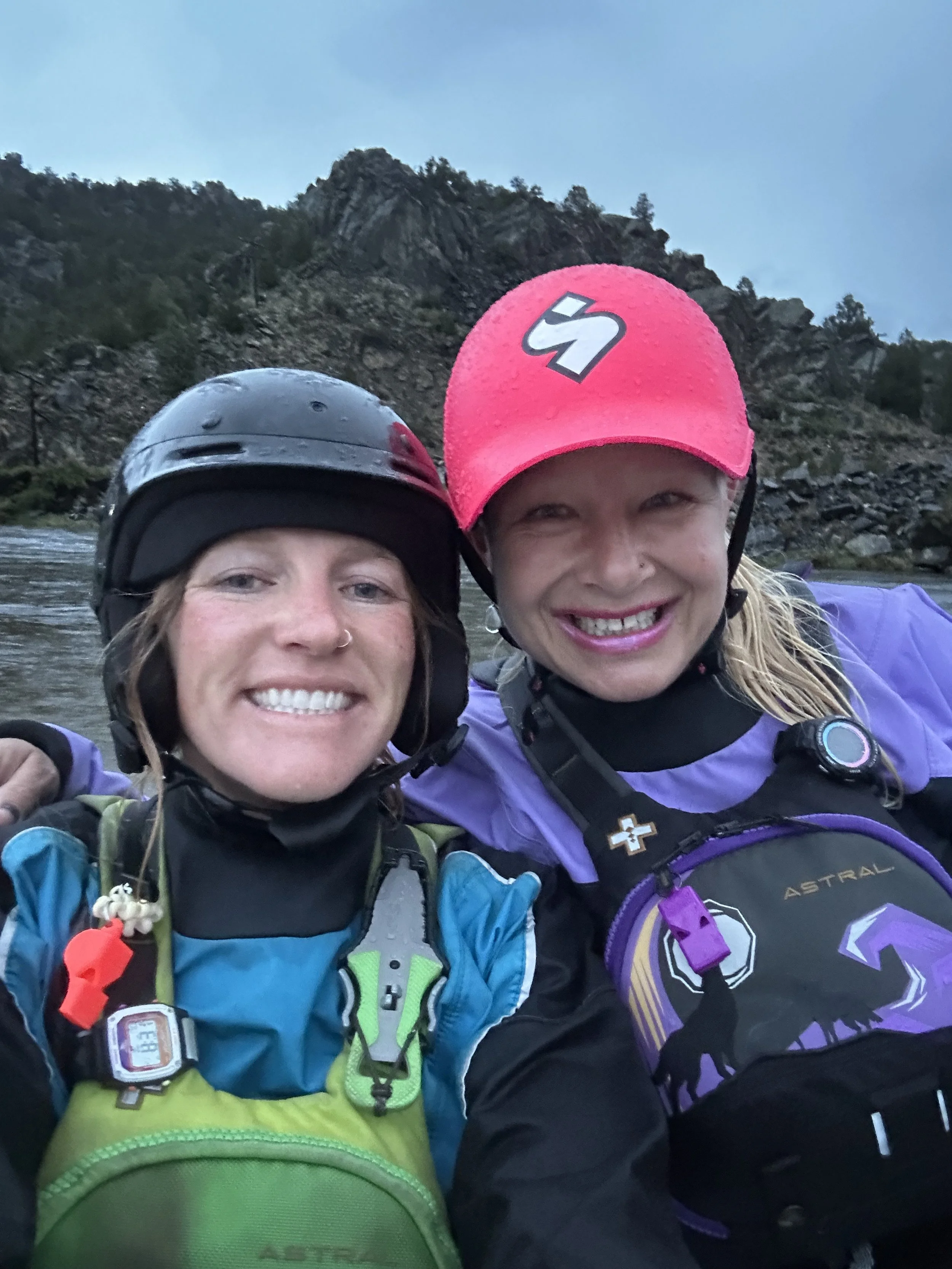 Two women wearing helmets and outdoor gear smiling for a selfie in front of a river and rocky hills.