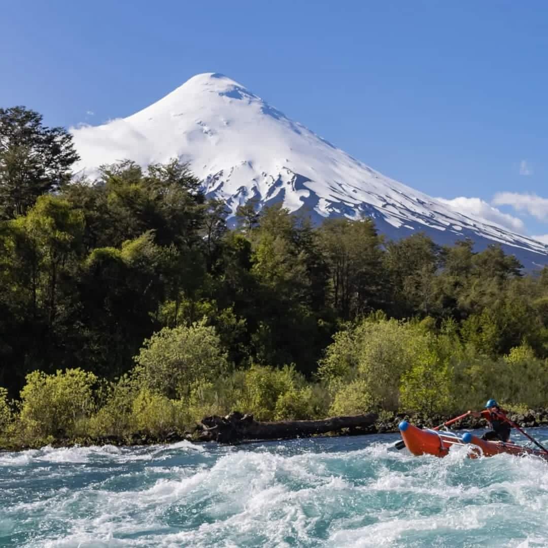 A boat in rushing water with lush green trees in the foreground and a snow-capped mountain in the background.