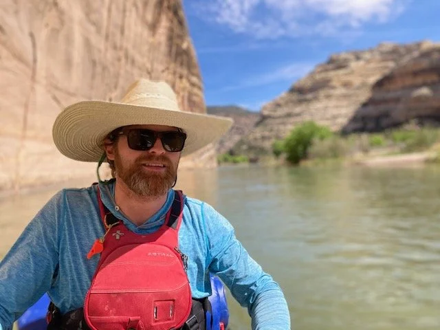 A man with a beard wearing sunglasses, a wide-brimmed hat, and a blue long-sleeve shirt, sitting in a boat on a river surrounded by rocky cliffs and trees under a blue sky.