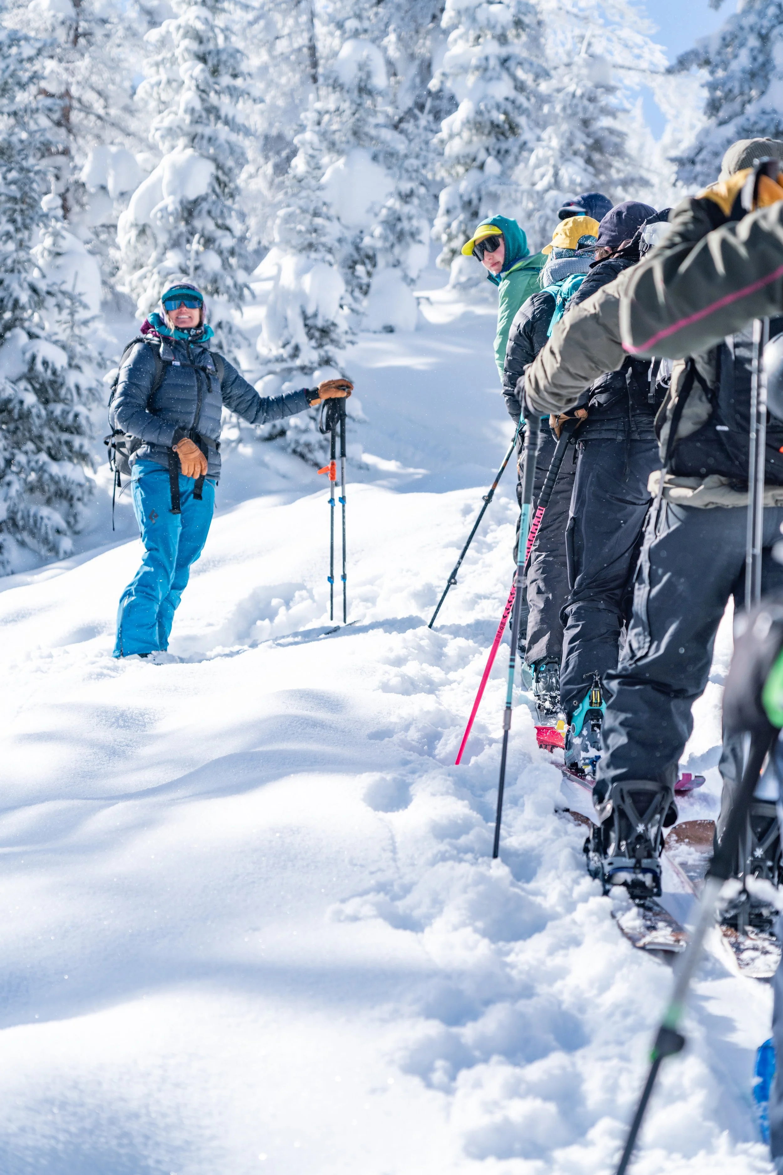 Group of people in winter clothing standing in a snow-covered forest, with one person facing the camera holding ski poles, as the others listen attentively.