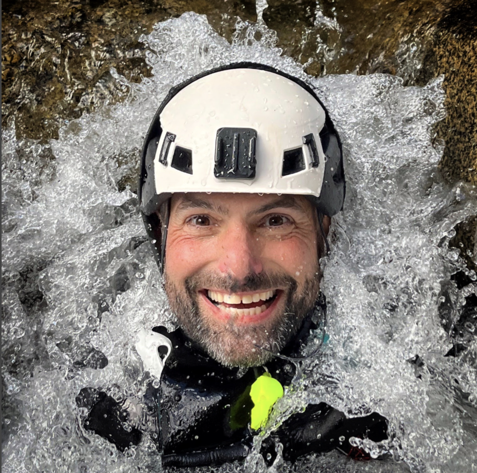 A man smiling while wearing a helmet in a river, surrounded by splashing water.