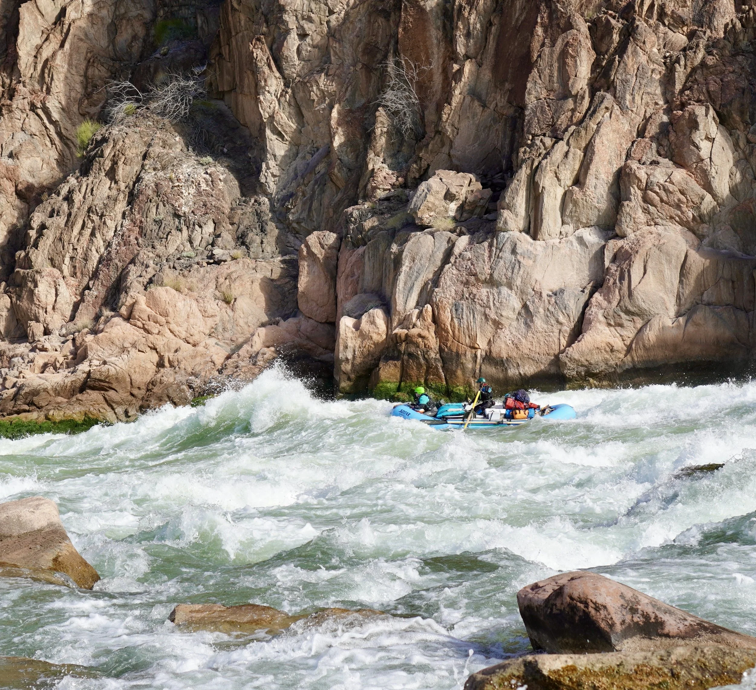 A group of people whitewater rafting on a rocky river with large boulders and steep, rugged cliffs in the background.