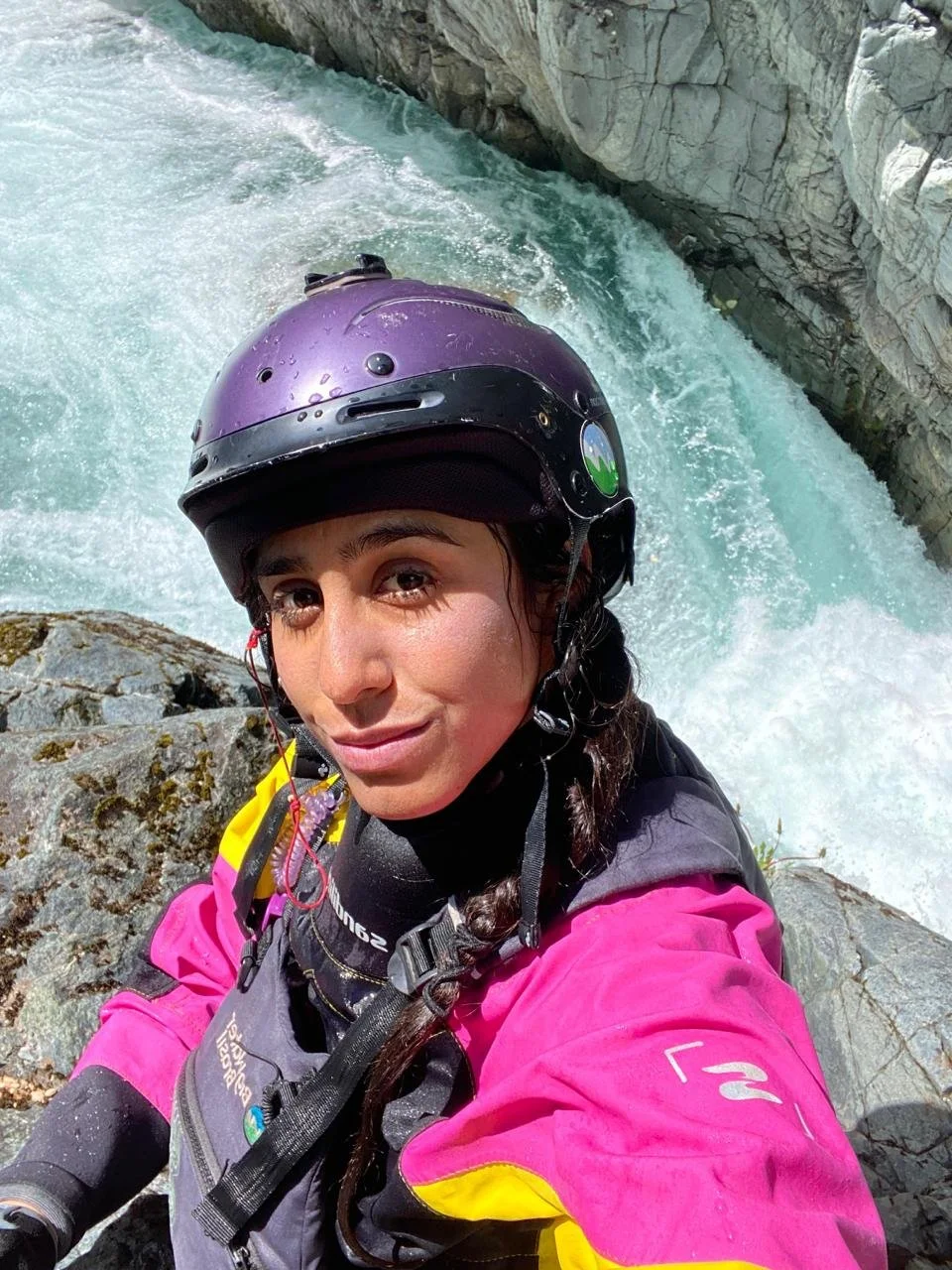 A woman in a pink and yellow waterproof jacket and a purple helmet taking a selfie near a fast-flowing river or waterfall with rocky cliffs in the background.