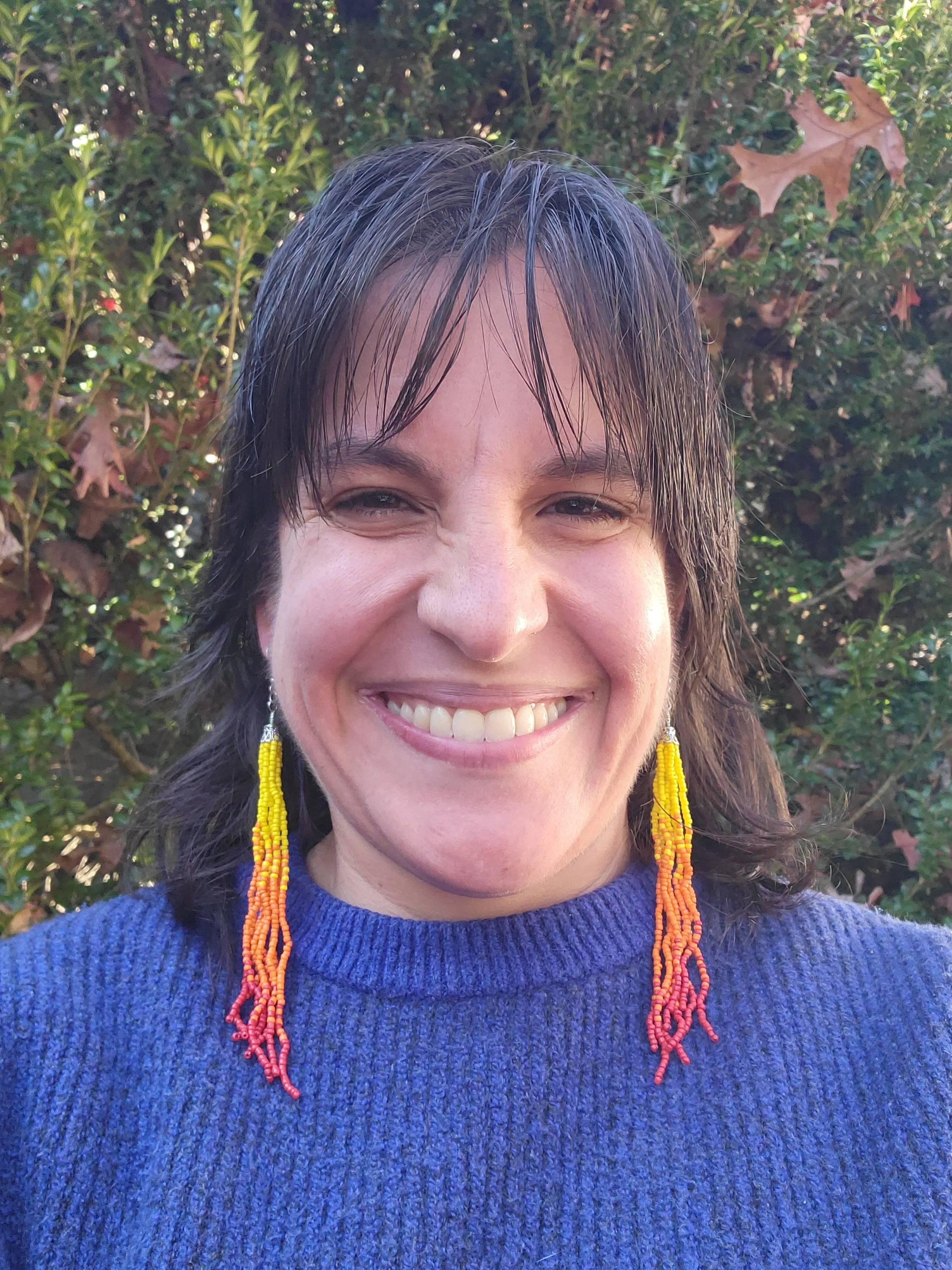 A woman smiling outdoors in front of green bushes, wearing a blue sweater and colorful bead earrings.