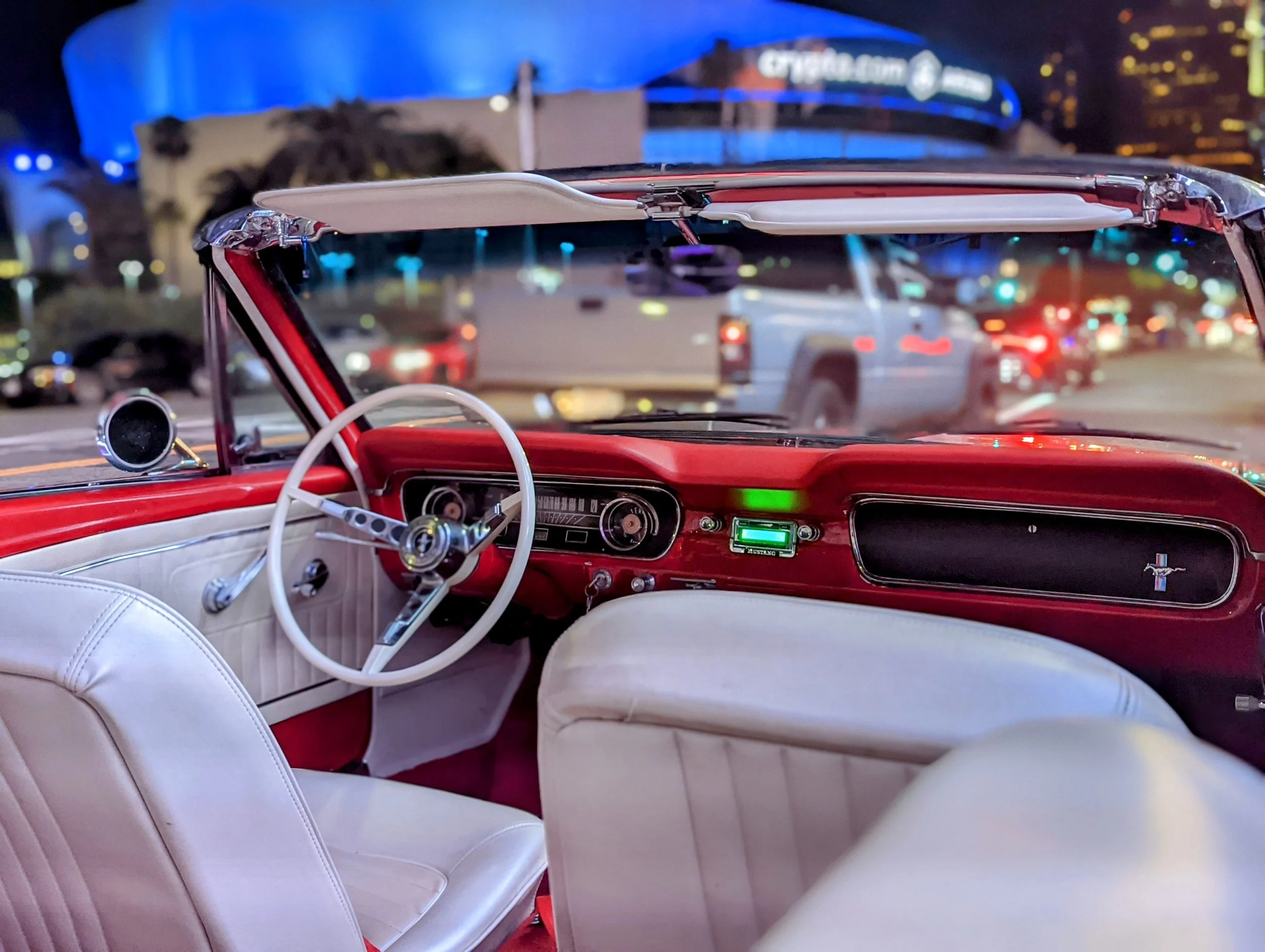 Interior of a vintage red and white convertible car, showing the dashboard, steering wheel, and front seats, with city lights and traffic visible through the windshield at night.