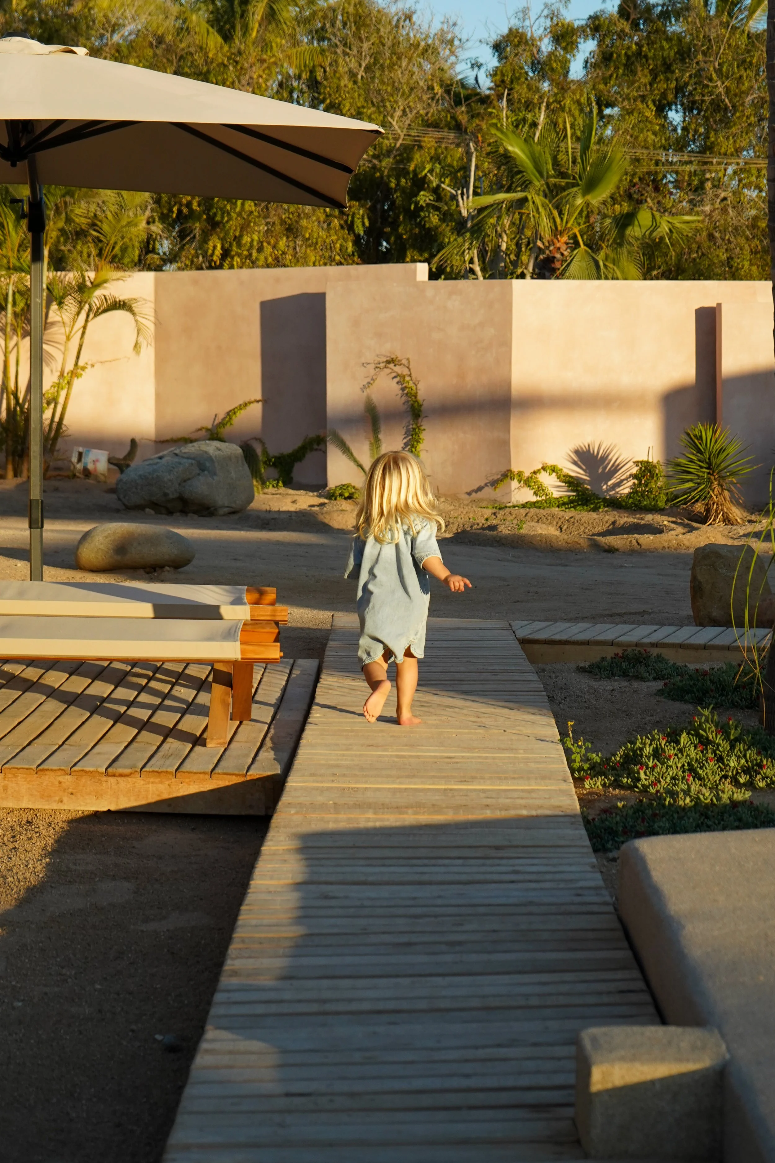 A young girl with blonde hair walking barefoot along a wooden pathway outdoors during golden hour, with a large umbrella, rocks, and desert plants in the background.